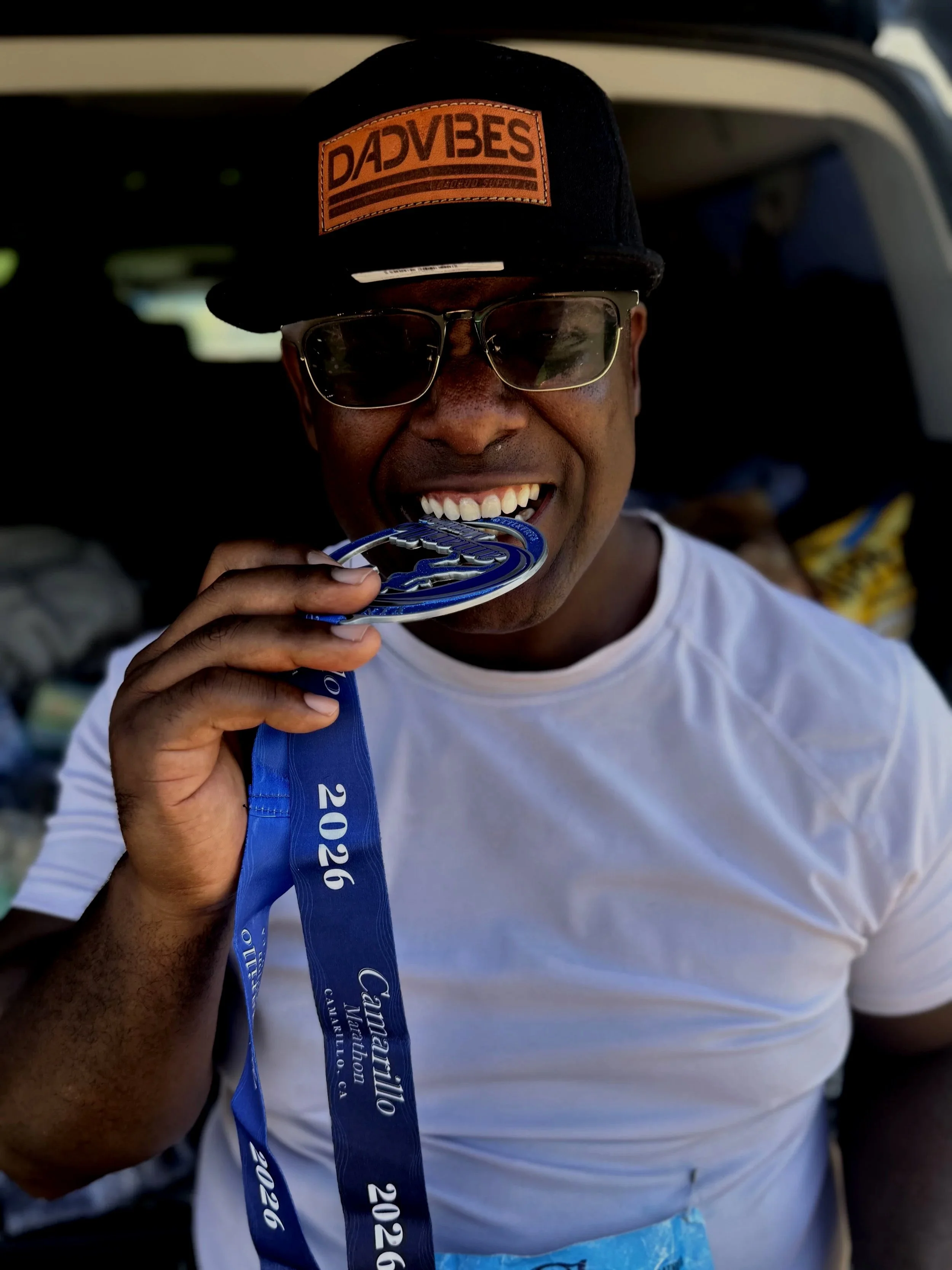 A man smiling while holding a blue medal in his hand. He is wearing sunglasses, a black cap with an orange patch that says 'DADVIES', and a white shirt. The background suggests an outdoor setting.