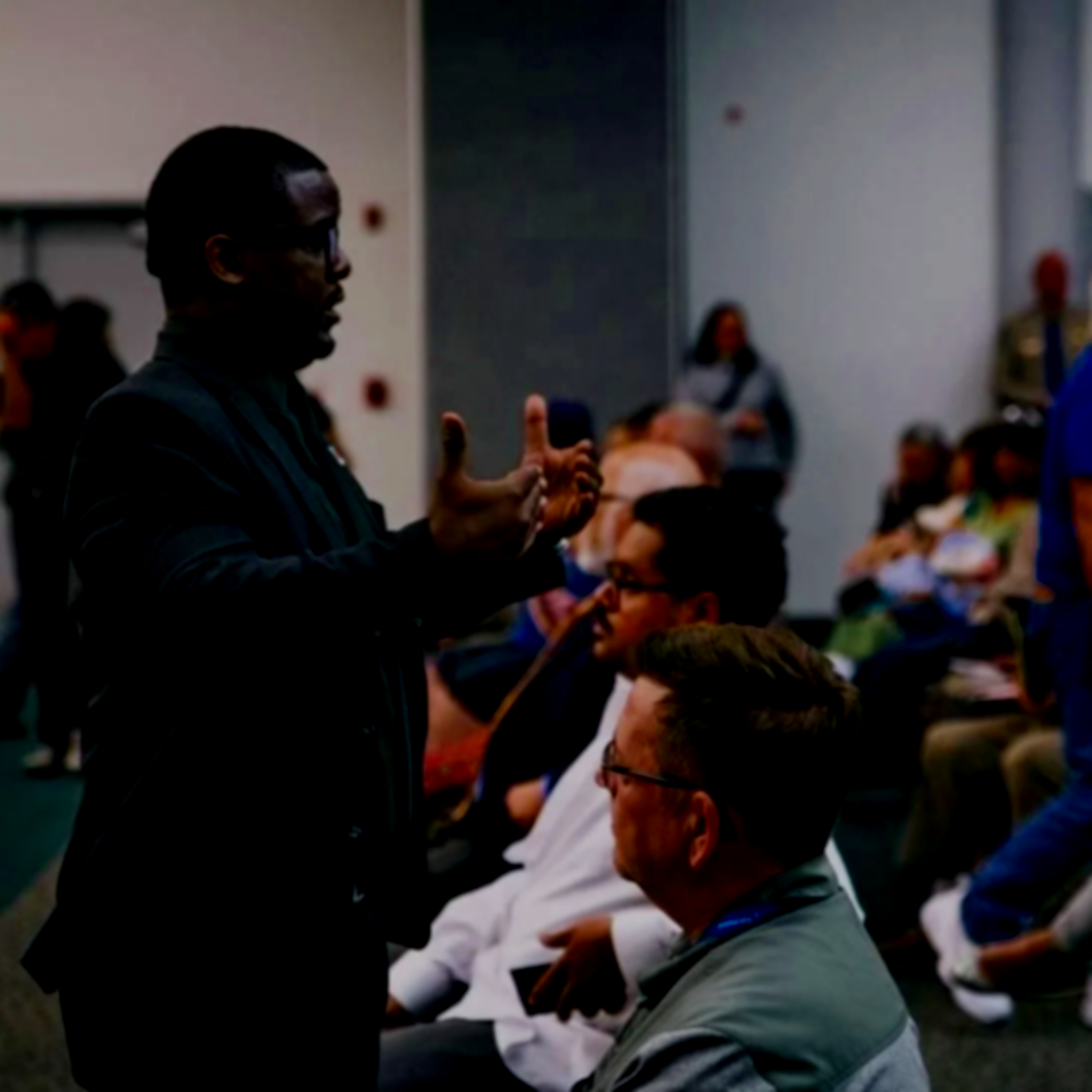 A man in a dark suit gestures with his hands while speaking to an audience. Several seated people listen, and a woman stands in the background in a room with a crowd.