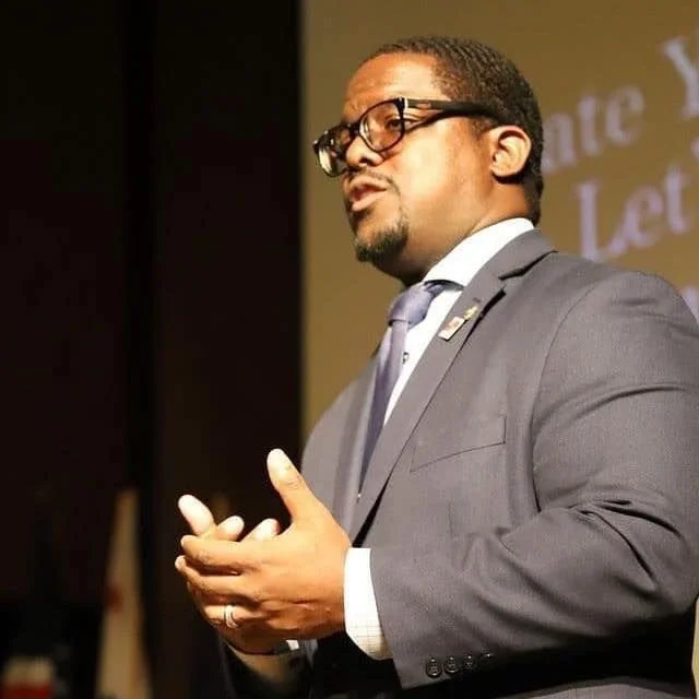 A man in a gray suit, white shirt, and purple tie giving a speech or presentation on stage, wearing glasses with short hair and a beard, with a banner or screen behind him.