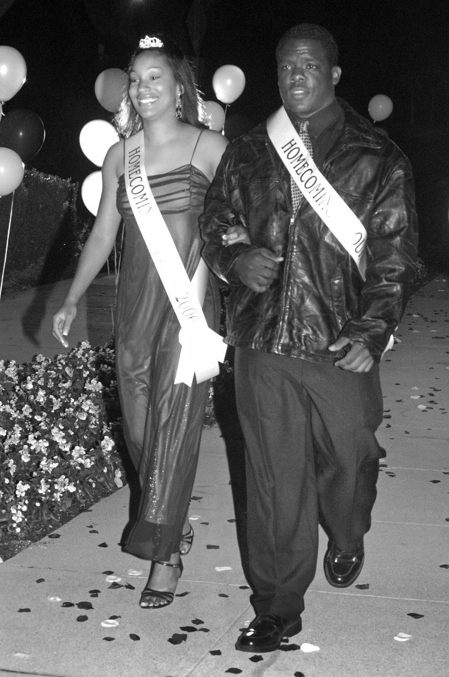 A young woman and a young man walking together at a celebration event, wearing sashes that say "Homecoming Queen" and "Homecoming King" respectively. The woman is smiling with a tiara, and the man is dressed in a leather jacket and formal pants, walking confidently amid balloons and flowers.
