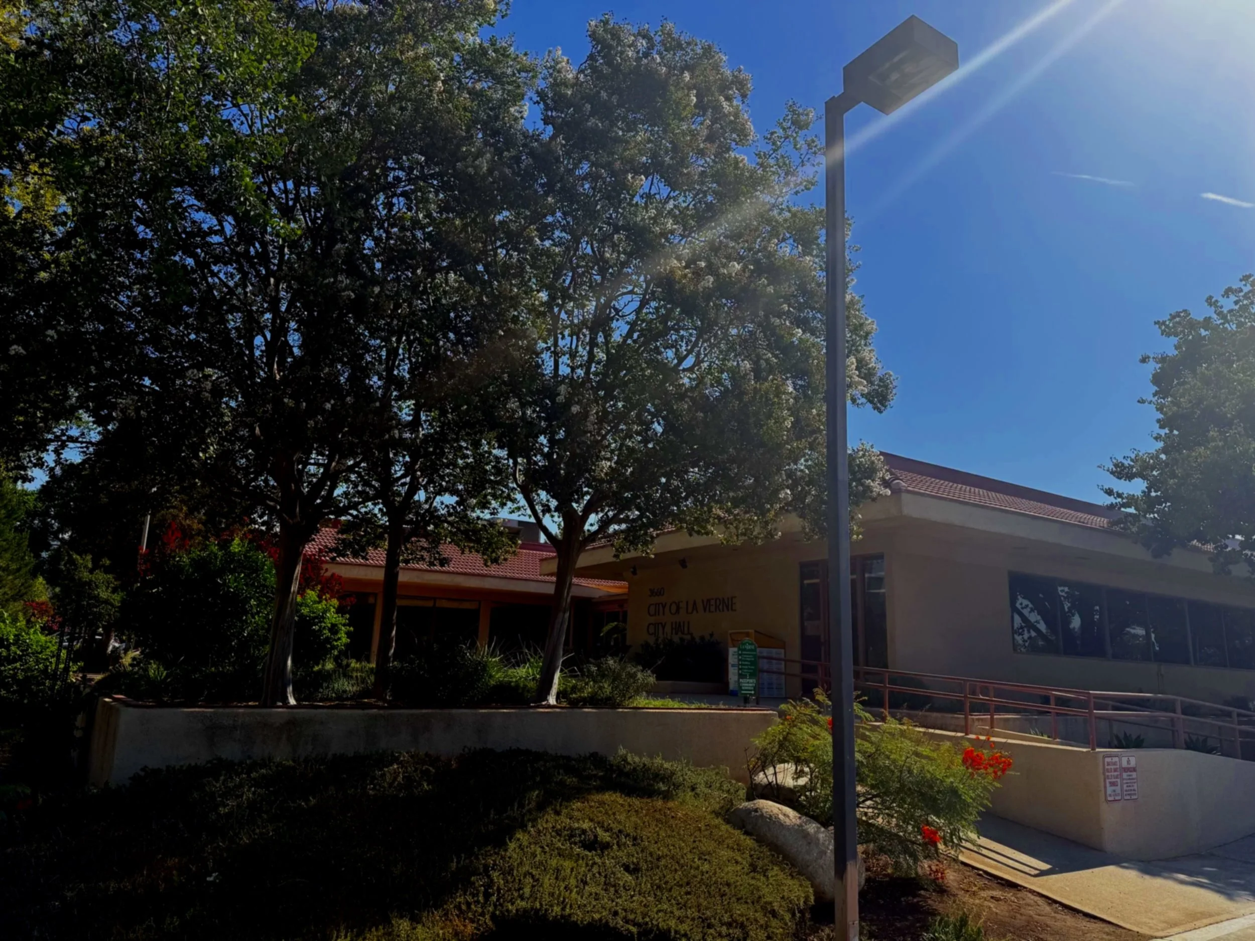A city hall building with a sign that reads 'City of La Verne City Hall' located in a landscaped area with trees and flowers, under a clear blue sky with the sun shining.