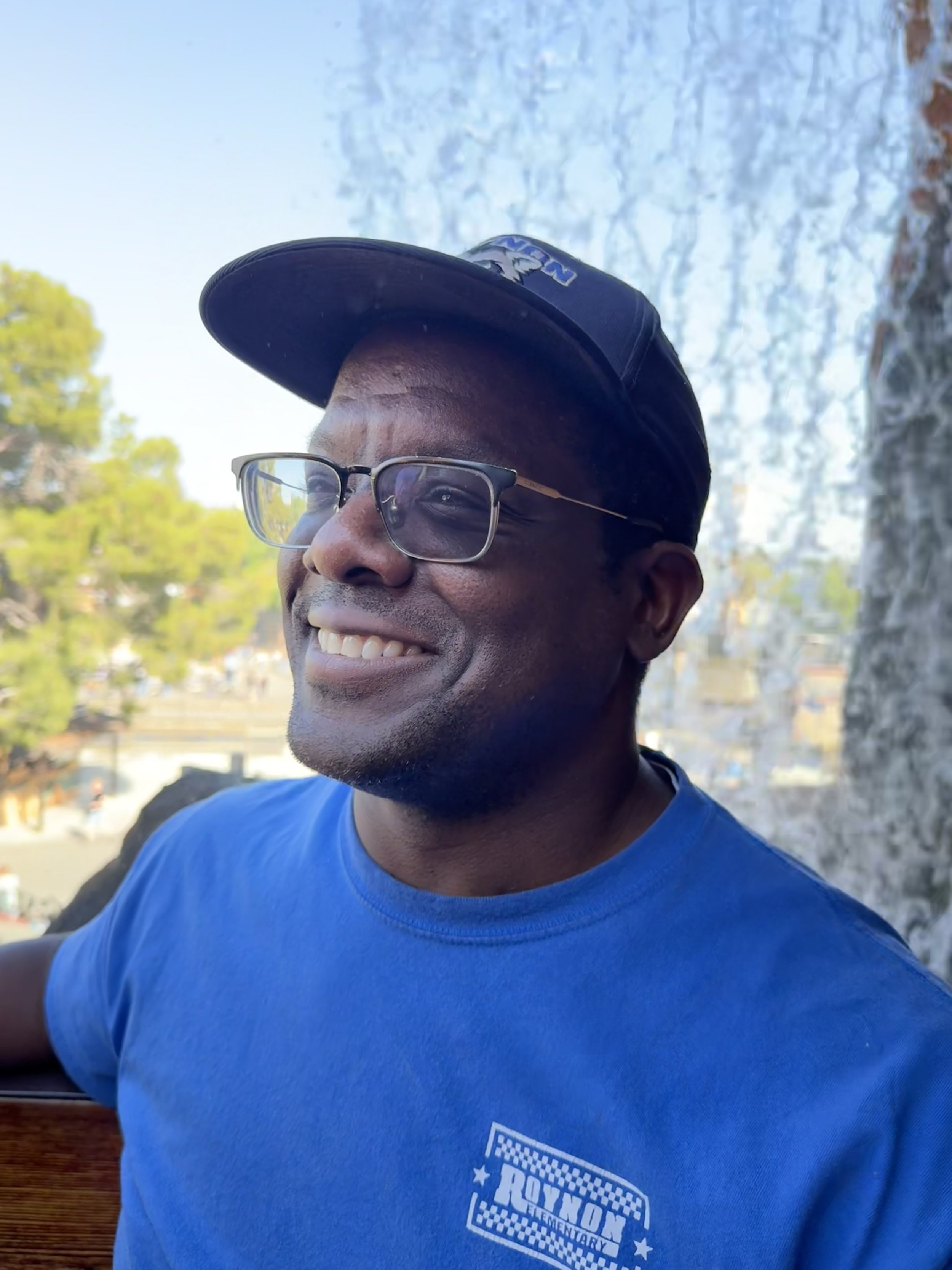 A man with glasses and a dark baseball cap smiling outdoors, wearing a blue T-shirt with a logo reading "Royal Elementary".
