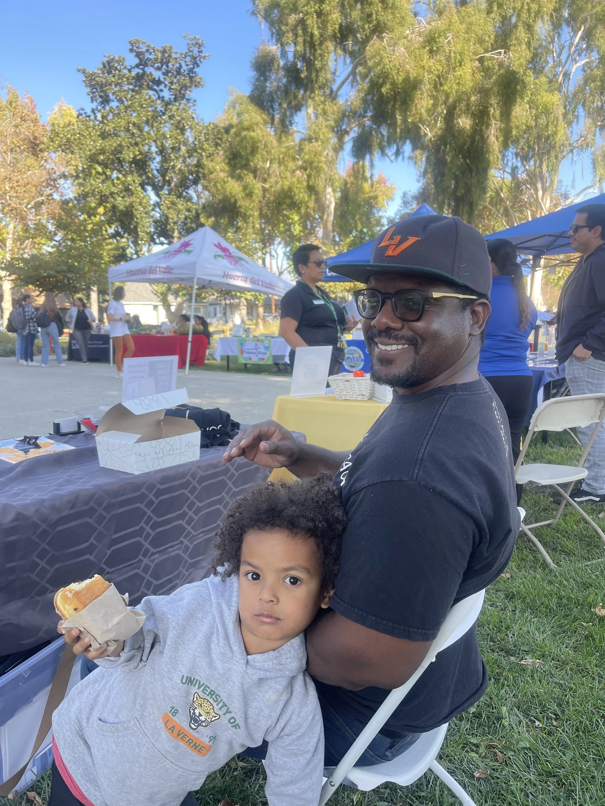 A man and a young girl sitting at an outdoor event, with tents and other people in the background, enjoying a sunny day.