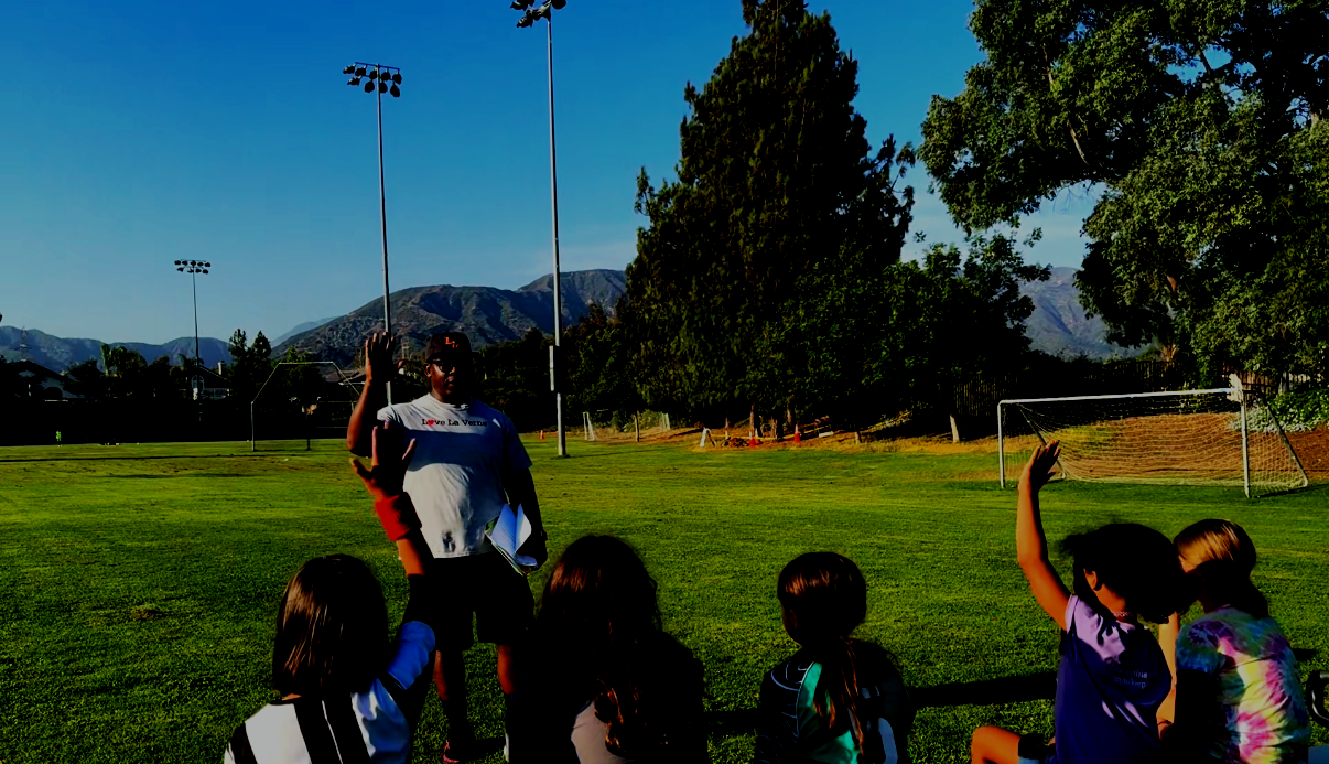 A coach on a soccer field giving instructions to children seated on the grass, with some raising their hands, on a sunny day with mountains and trees in the background.