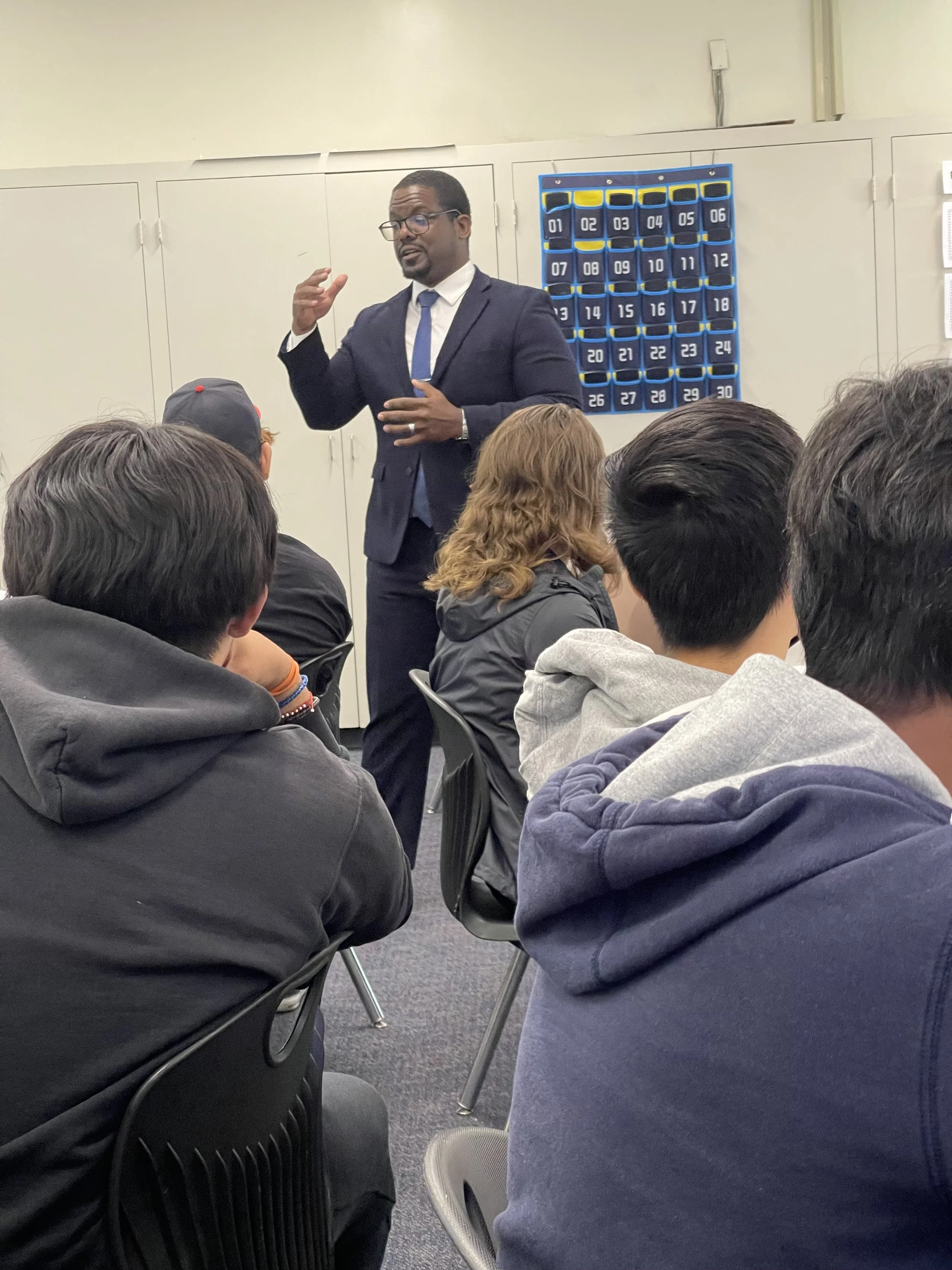 A man in a suit and tie giving a presentation to a group of students in a classroom. The students are seated and listening, with some wearing hoodies and caps. There is a blue pocket chart on the wall with numbered pockets, and cabinets in the background.