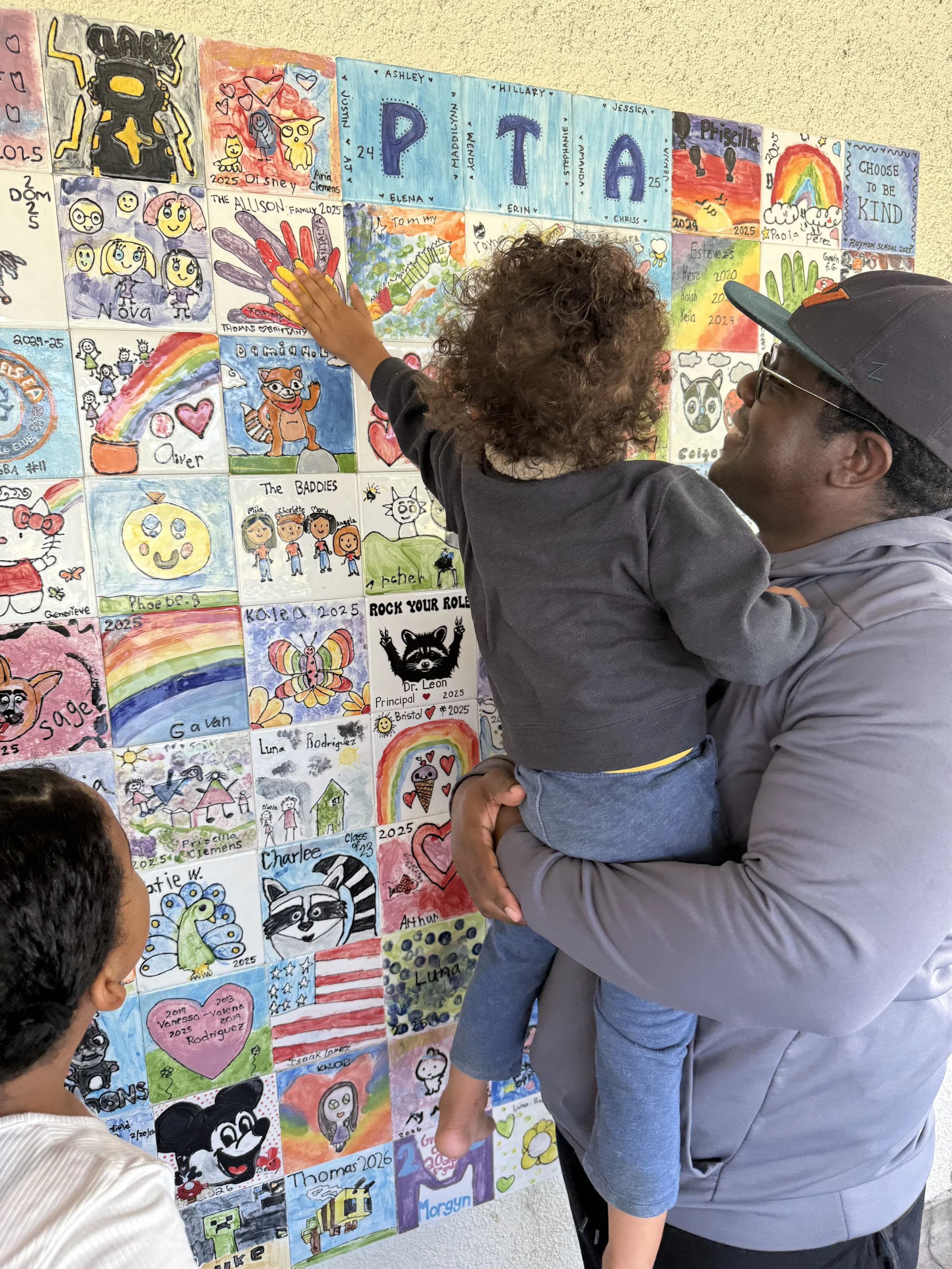 A man holding a child in front of a wall covered with colorful drawings and paintings, most of which appear to be children's artwork, including rainbows, characters, and various designs.