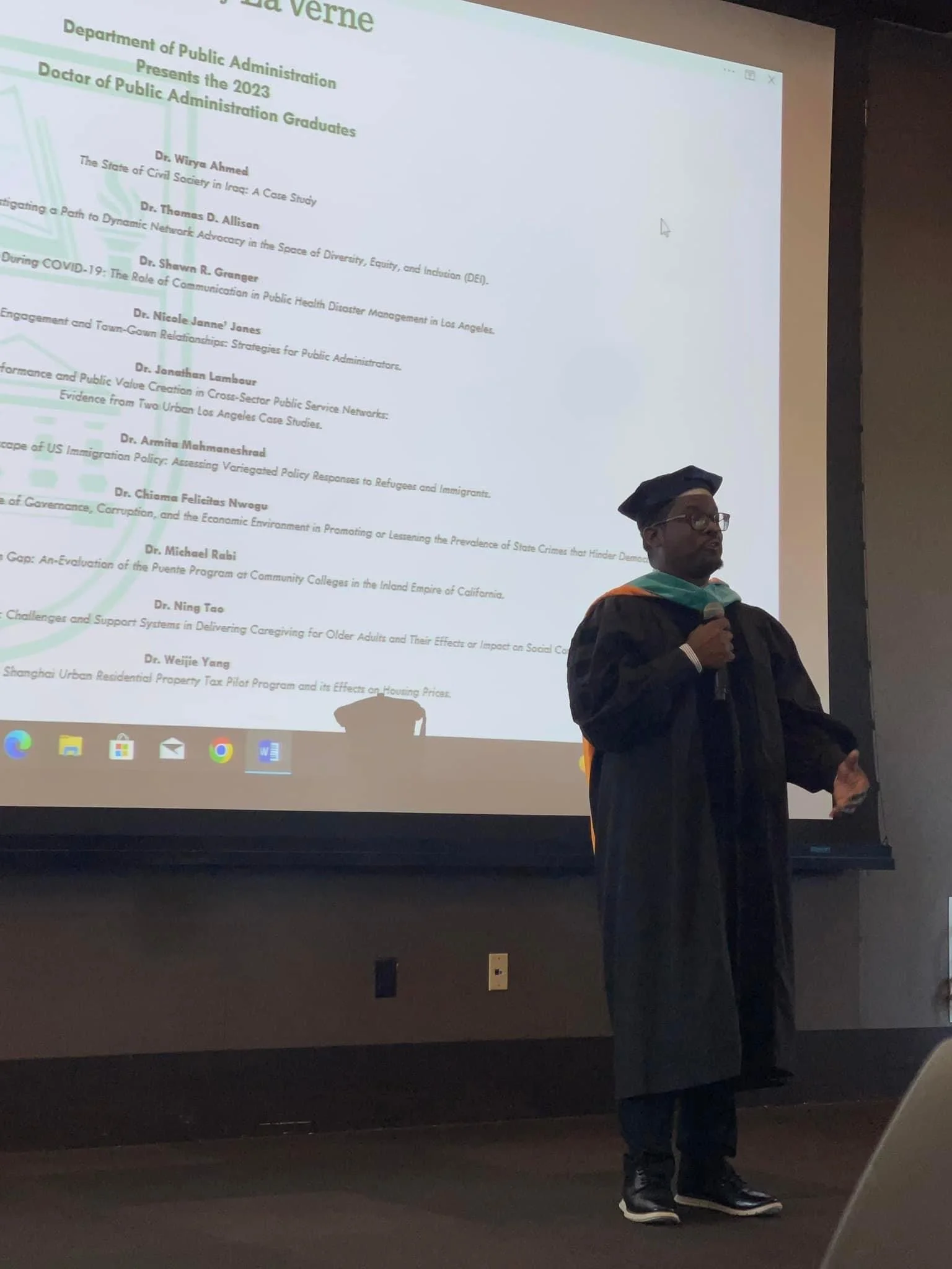 A person in academic regalia standing in front of a projection screen at a graduation ceremony.