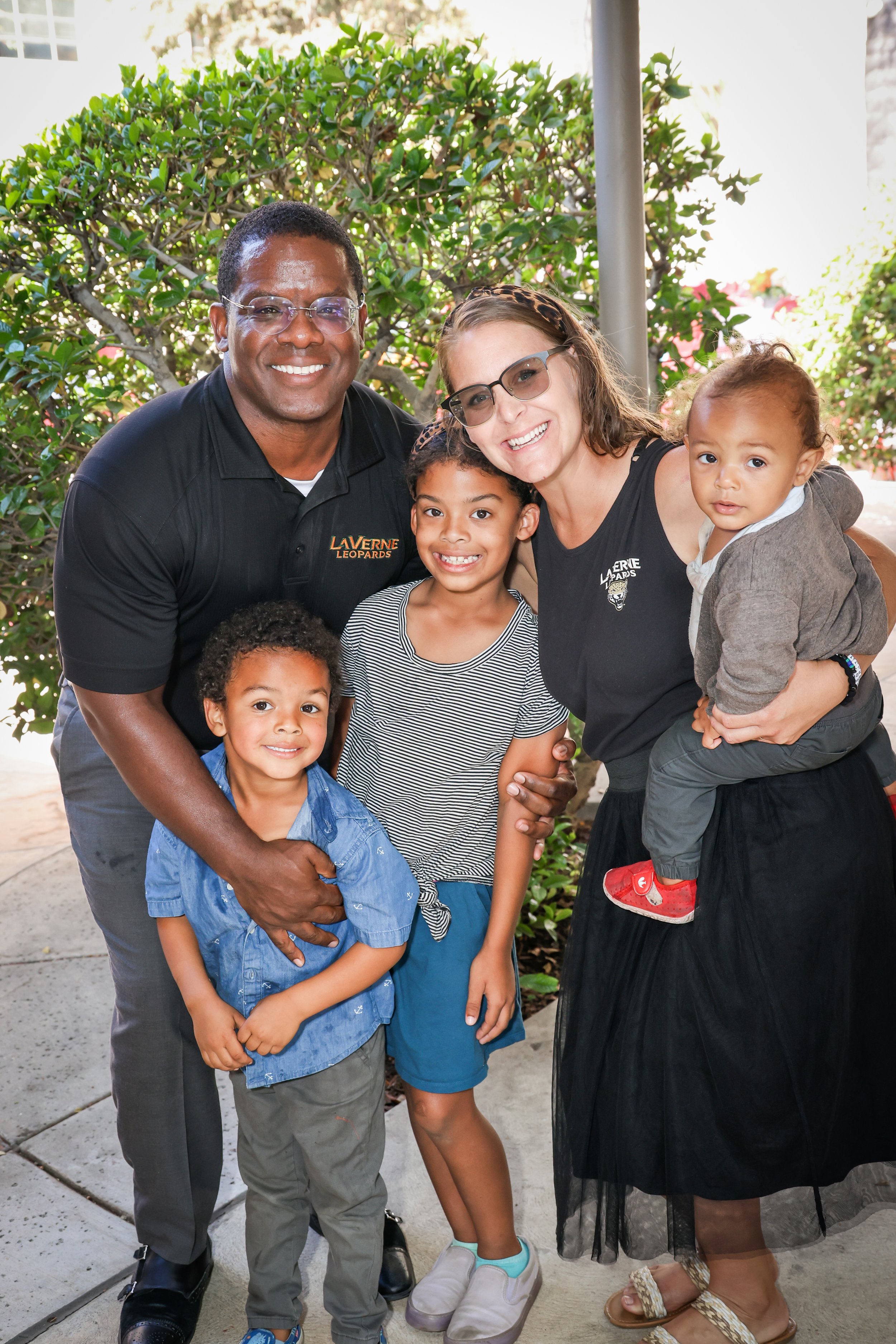 A diverse group of four children and two adults smiling and posing for a photo outdoors. The adults are a man in a black polo shirt and a woman in black glasses and a black dress, with the children standing close by. The background includes green shrubs and a light-colored building.