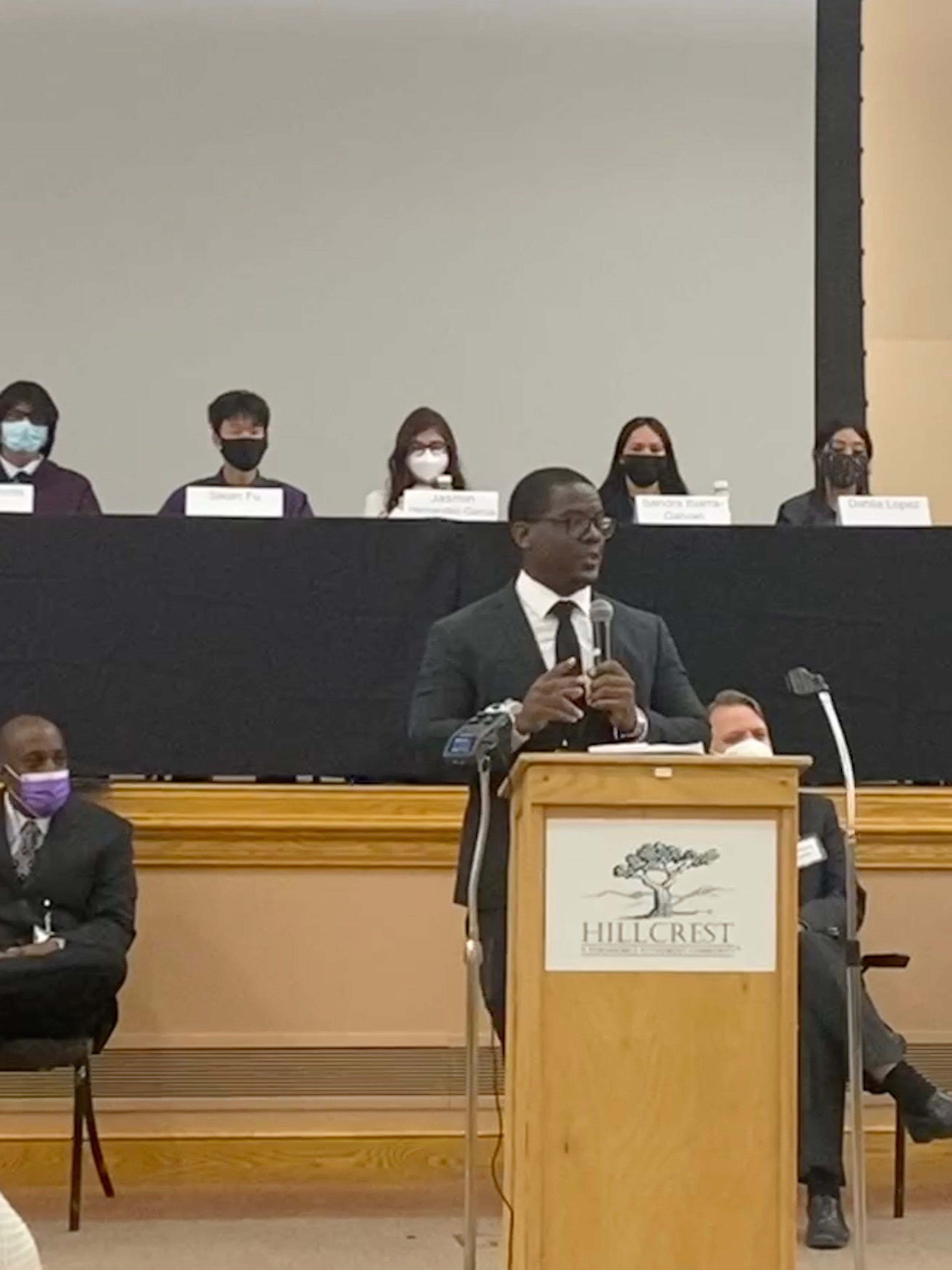 A man in a suit and tie speaking at a podium with a microphone, with a logo that says 'Hillcrest' and a tree on it, in front of an audience. Behind him, five people are seated at a long table, all wearing masks, with name tags in front of them.