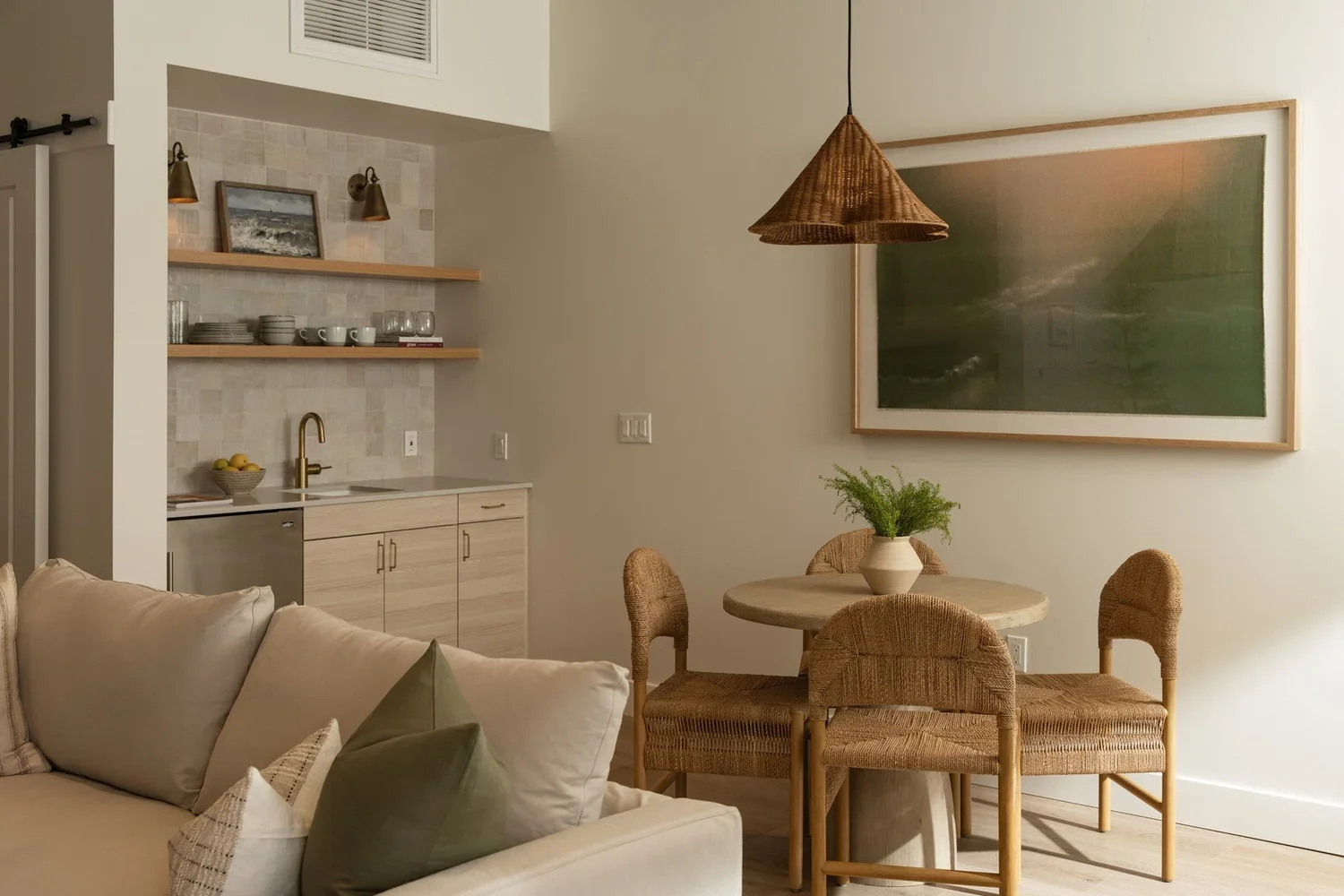 The image shows a cozy living and dining area with a soft beige couch, a round wooden table with four woven chairs, and a small kitchenette with open shelves, a sink, and a countertop with a bowl of lemons. A large framed artwork hangs on the wall, and a woven pendant light hangs above the dining table. There is a green plant in a white vase on the table.