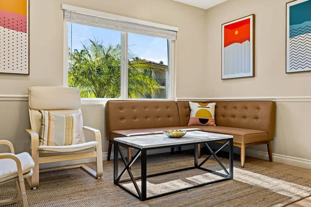 Living room with beige walls, window with blinds, tan corner sofa, white armchair with striped pillow, framed abstract art on walls, coffee table with decorative bowl, and tropical trees outside window.