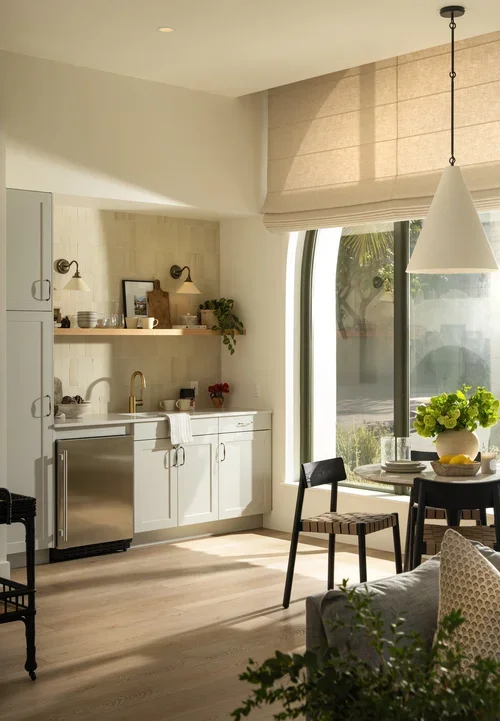 Bright kitchen and dining area with white cabinets, a small sink, open shelves, and a large window with a roman shade. There is a round dining table with black chairs and a hanging white pendant light. A vase with green hydrangeas decorates the table, and a couch with pillows is partially visible in the foreground.