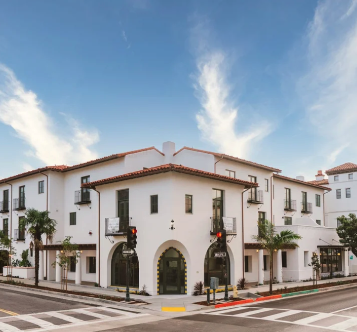 White multi-story residential building with arched entrances, small balconies, and palm trees along the street. Traffic lights and crosswalks are visible in the foreground, under a blue sky with scattered clouds.