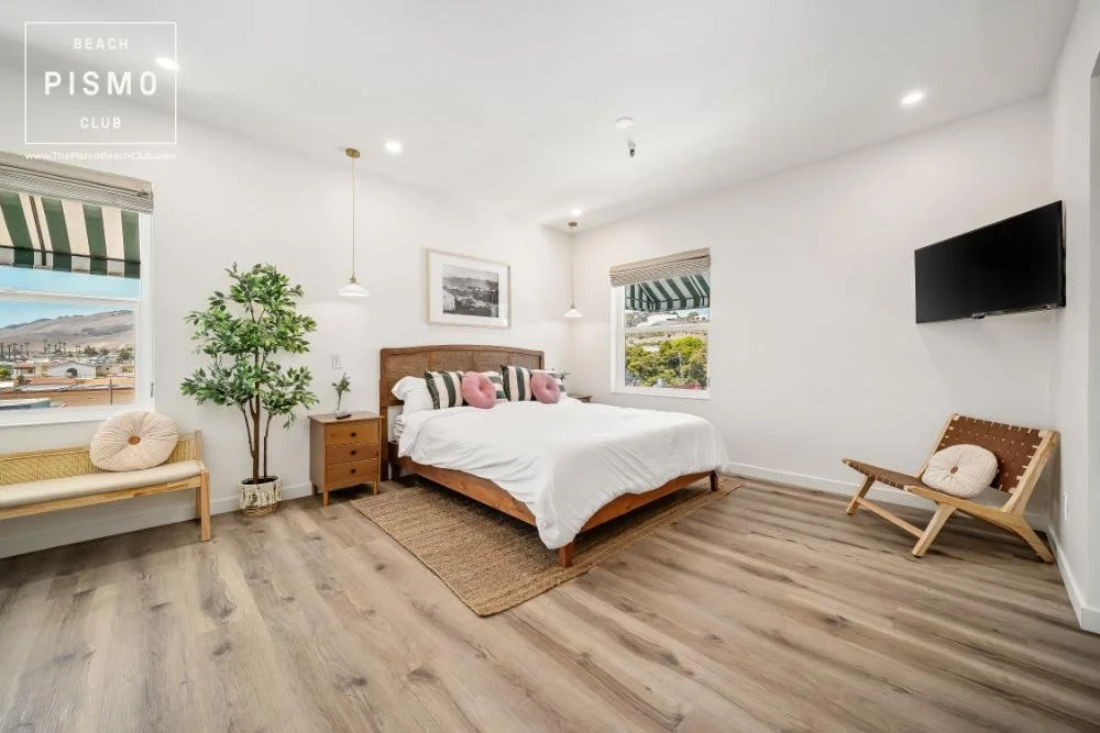 A bright bedroom with a wooden bed frame, white bedding, and pink and striped pillows. There are two windows with green and white striped blinds, a potted plant, a wooden bench with a decorative cushion, a wall-mounted flat-screen TV, and a small wooden chair with a cushion. The room has light-colored flooring and white walls.