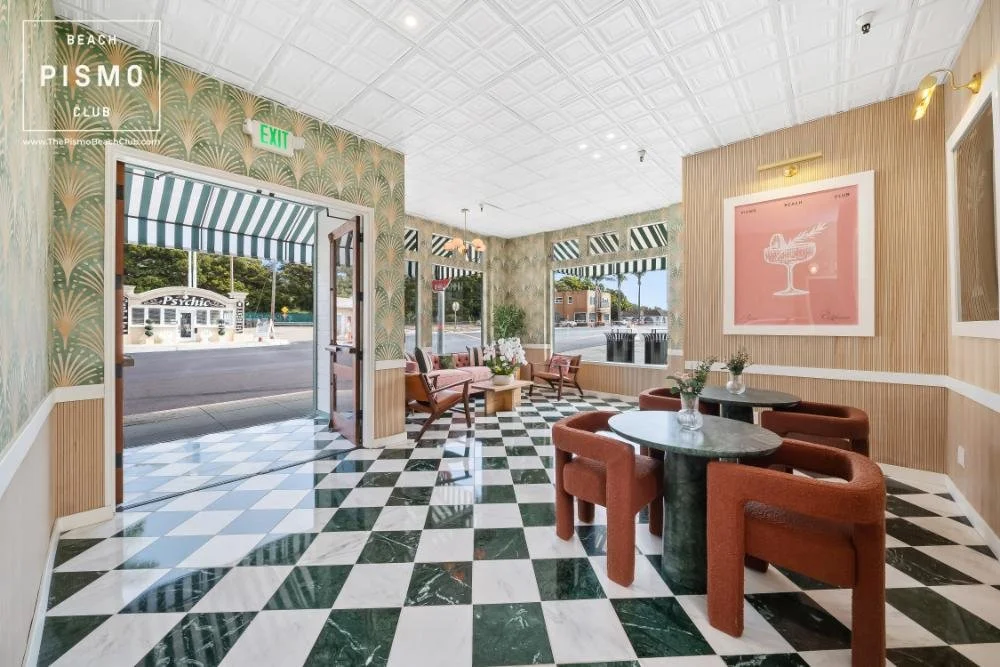 Interior of a stylish cafe or lounge with checkered black and white marble flooring, patterned wallpaper, and modern furniture including round tables with brown upholstered chairs and a seating area with armchairs. Large windows provide natural light and outdoor views. Decor includes framed pink artwork and flower vases on tables.