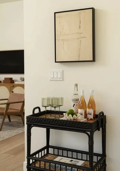 A black rattan serving cart with drinks and glasses, a framed abstract artwork, and a light switch on a white wall in a modern living space.