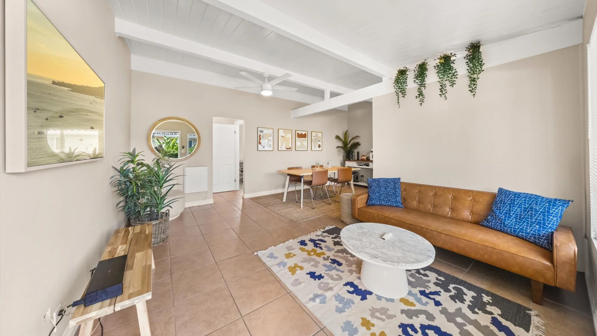 Living room with tan leather couch, blue pillows, wooden dining table, beige walls, white ceiling, indoor plants, wall art, sliding glass door, tiled floor, and a round white coffee table.