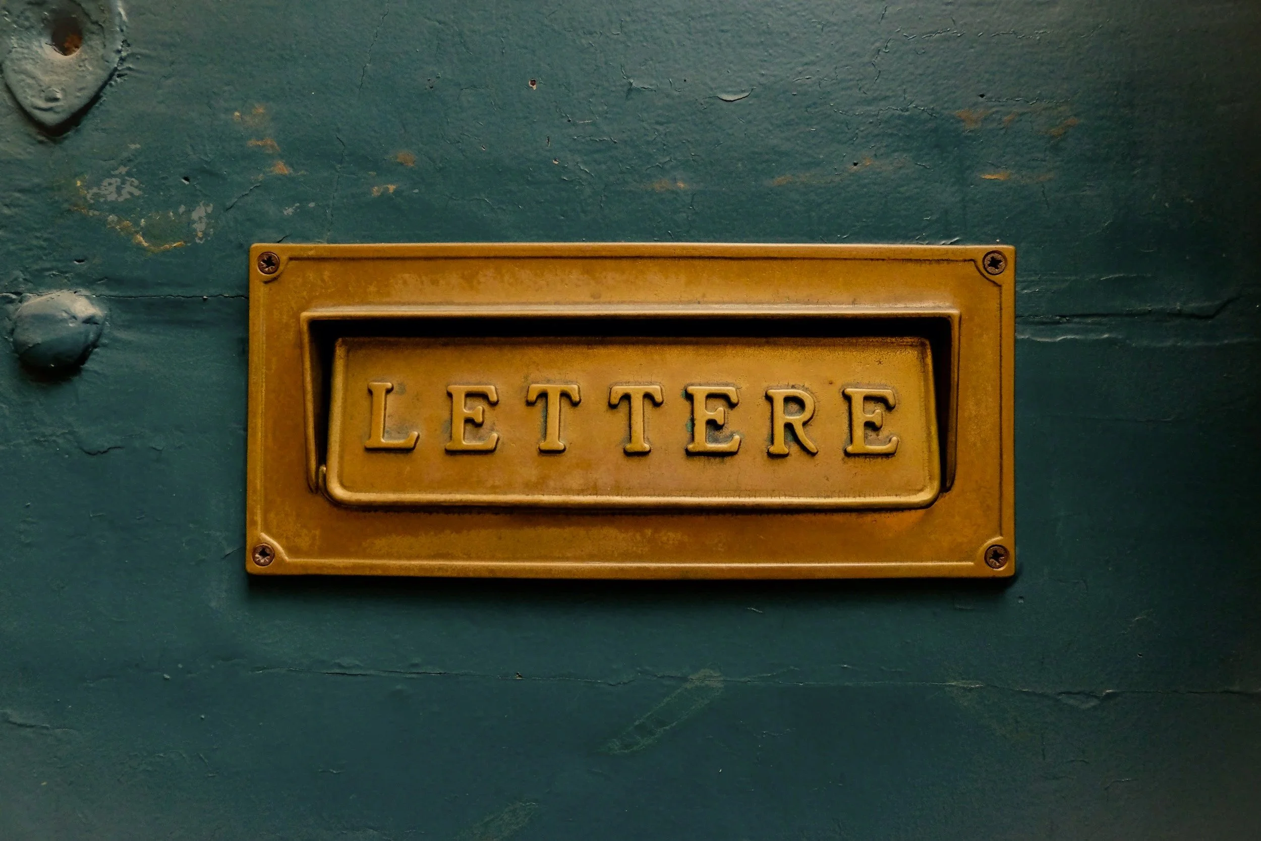 A vintage brass mailbox with the word 'LETTERS' inscribed on it, mounted on a weathered green wall.