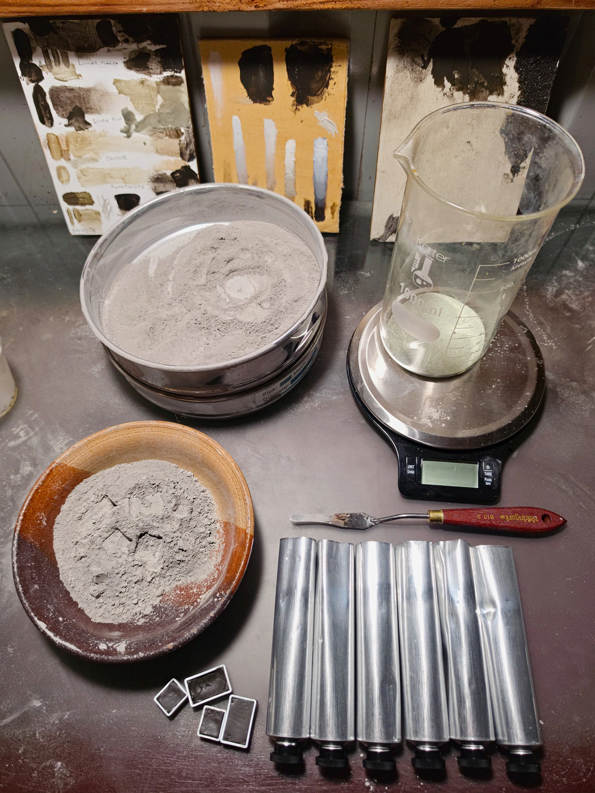 A laboratory workspace with a round bowl of grey powder, a digital scale, a measuring beaker with a liquid, and various metal molds and tools on a dark surface.