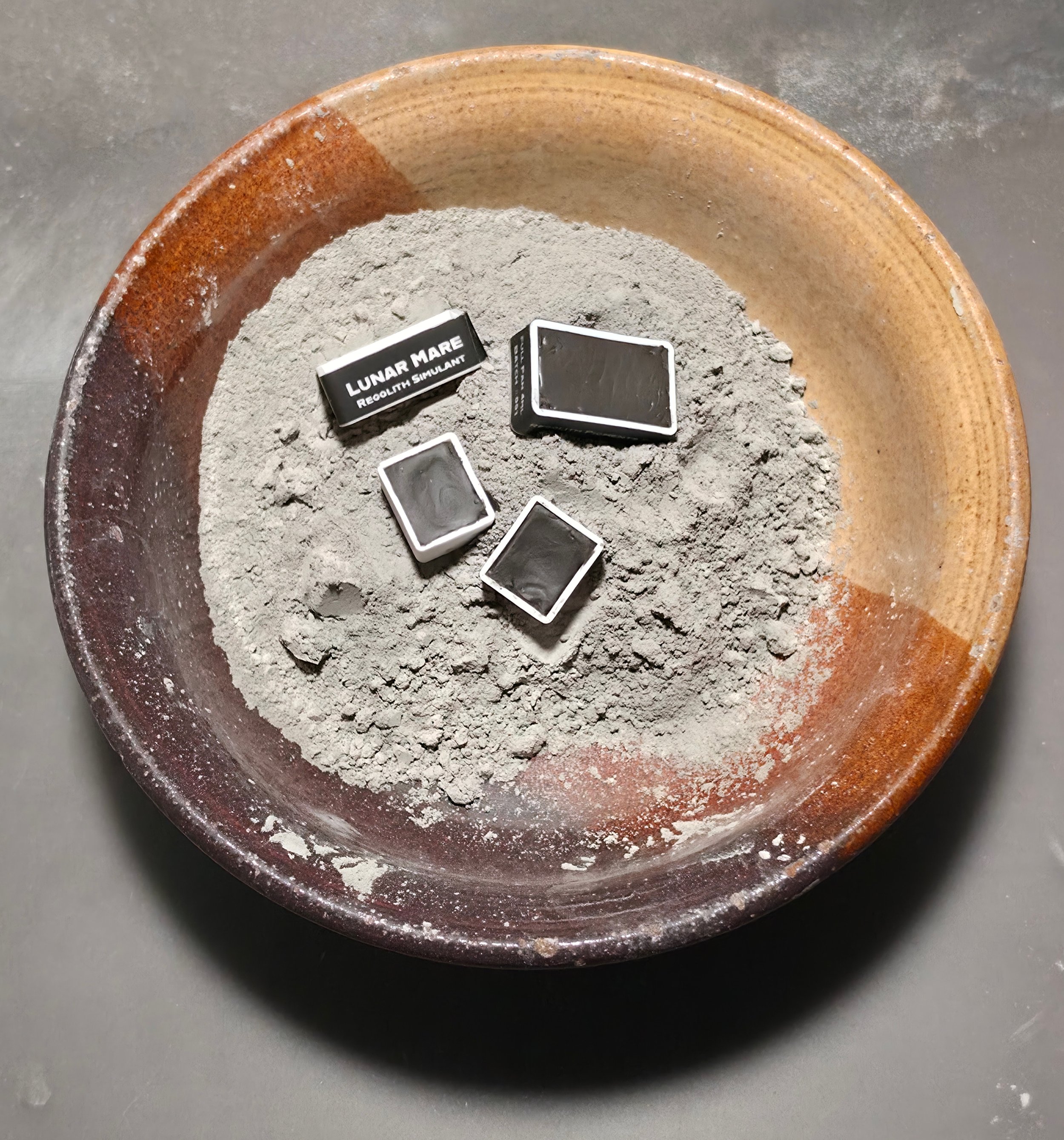 A round ceramic bowl filled with gray powder, with small rectangular black and white boxes labeled "Lunar Mare Reolithic Simulant" resting on top of the powder.