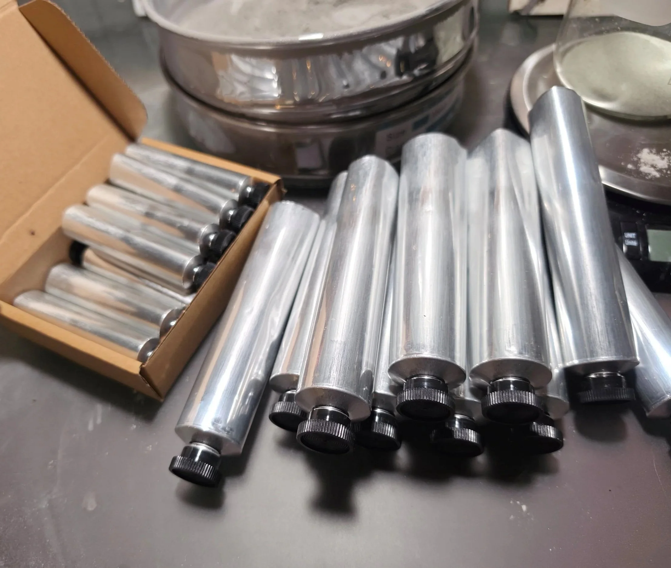 A bunch of small, shiny aluminum tubes with black caps at one end, lying on a dark surface, with a cardboard box of similar tubes and some other kitchen utensils nearby.