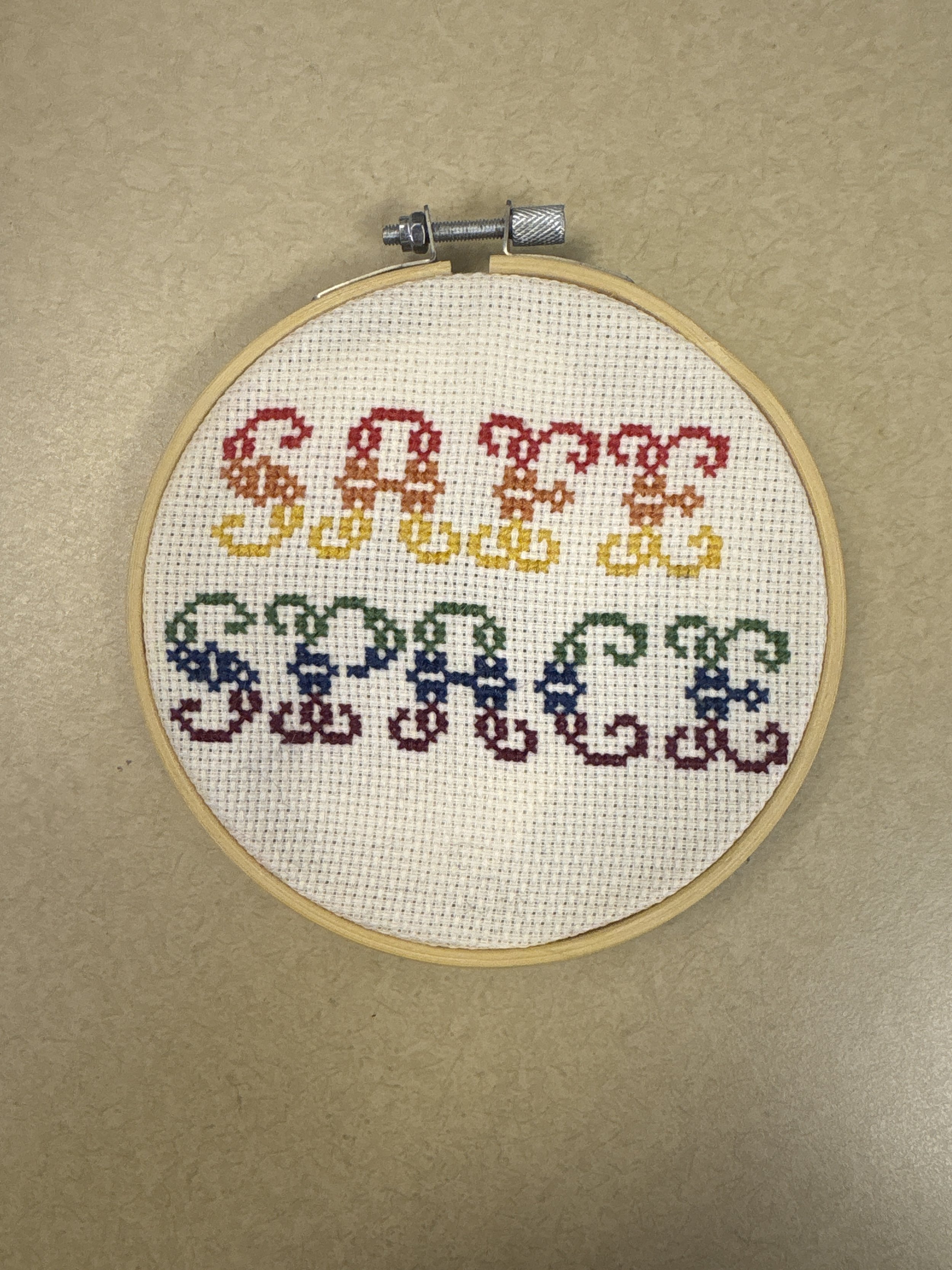 Cross-stitched word 'SAFE SPACE' in rainbow colors inside a wooden embroidery hoop on a beige surface.