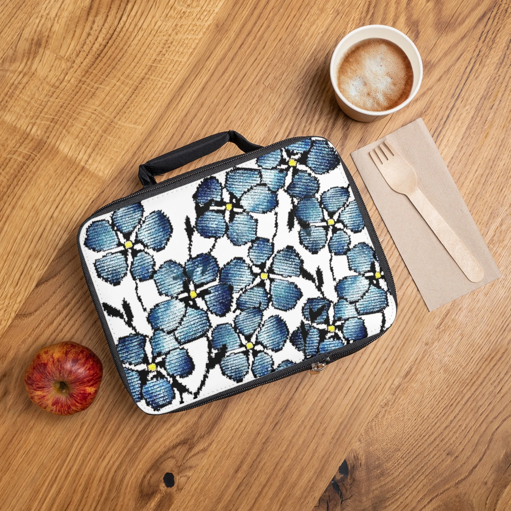 A wooden table with a cup of frothy coffee, a floral patterned lunchbox, a fork on a napkin, and a red apple.