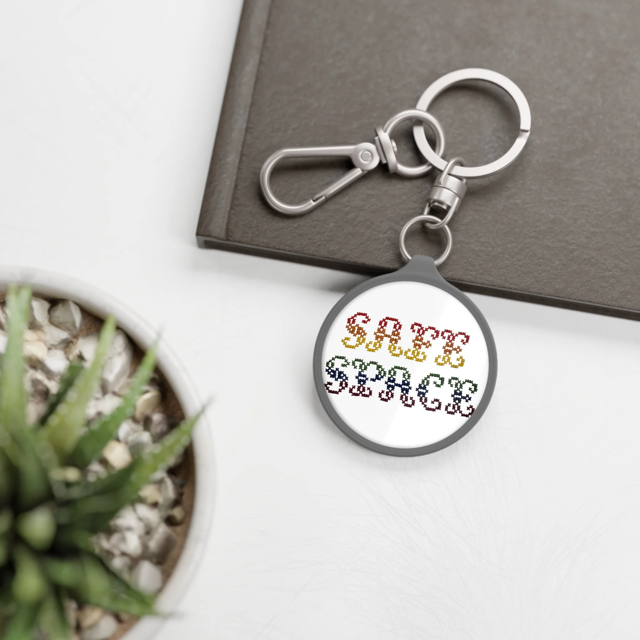 Close-up of a metal carabiner and keyring on a grey surface with rainbow-colored cross-stitch that spells out 'Safe Space'.