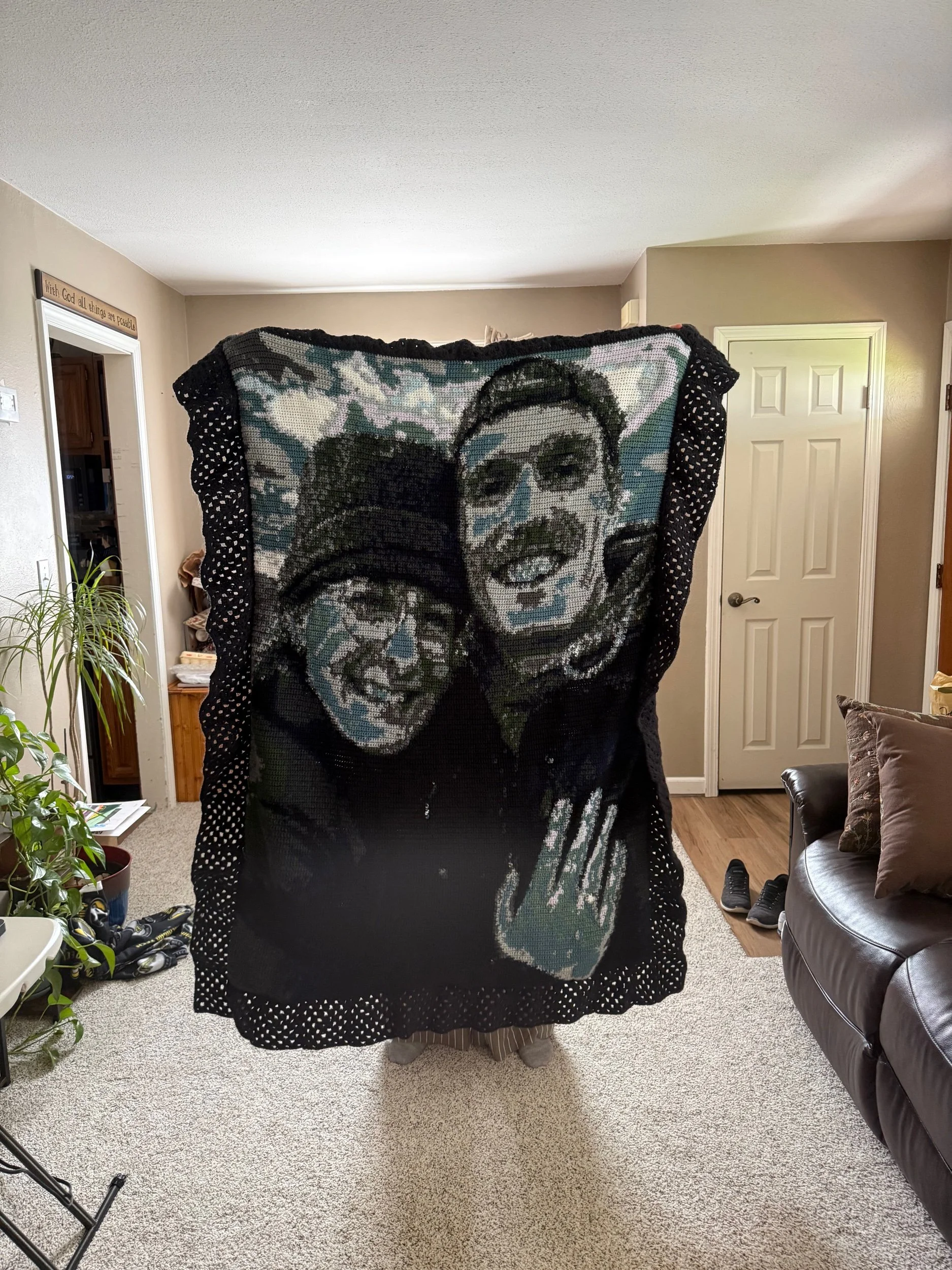 Person standing indoors holding a large blanket with a portrait of two people on it. The blanket features a colorful, pixelated depiction of a smiling man and woman wearing glasses.