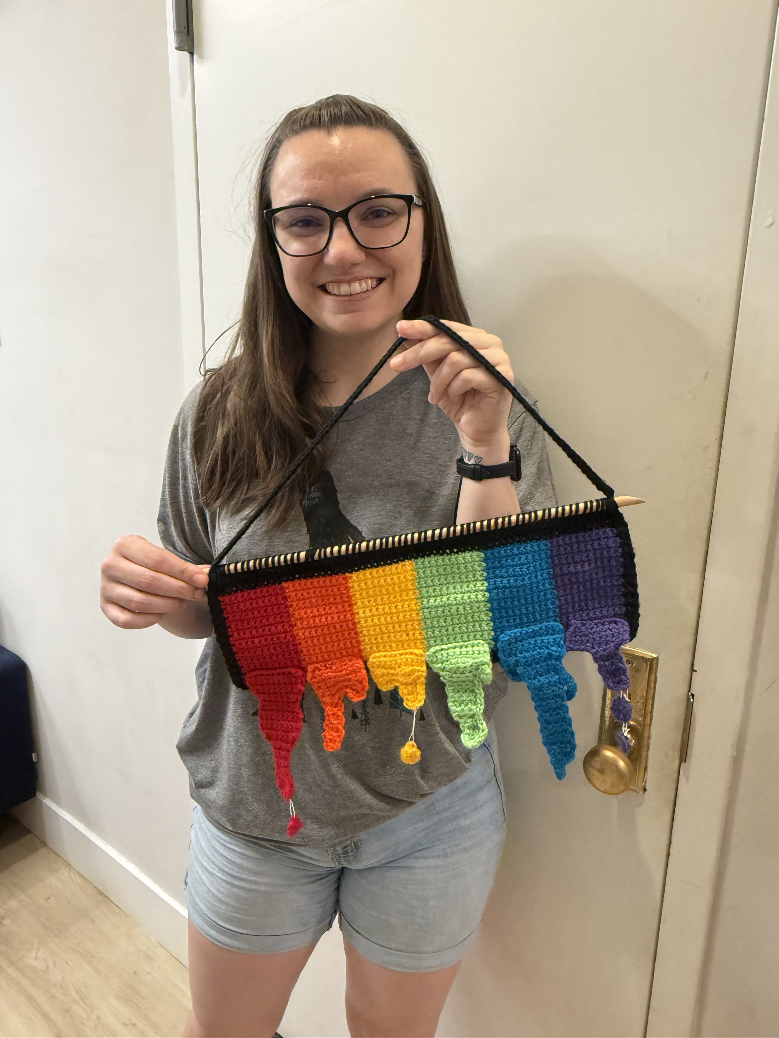 Rebecca smiling and holding up a rainbow-colored woven wall hanging.