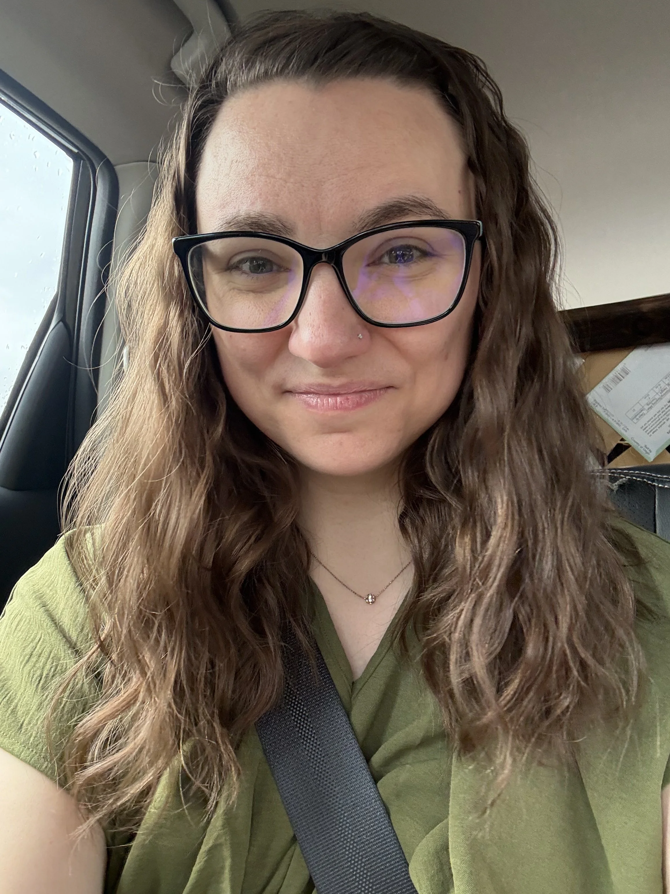 A woman with long, wavy brown hair wearing glasses and a green shirt, sitting in a car with a seatbelt on. She has a nose piercing and a small necklace, and there are packages or boxes visible in the background.