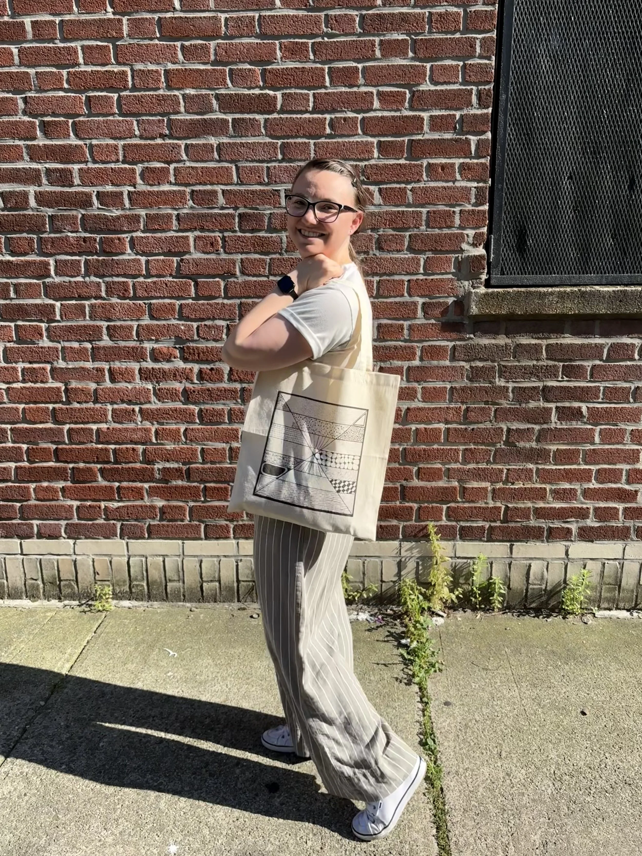 Rebecca holding a beige tote bag stands against a brick wall in sunlight, wearing a light-colored t-shirt and striped pants.