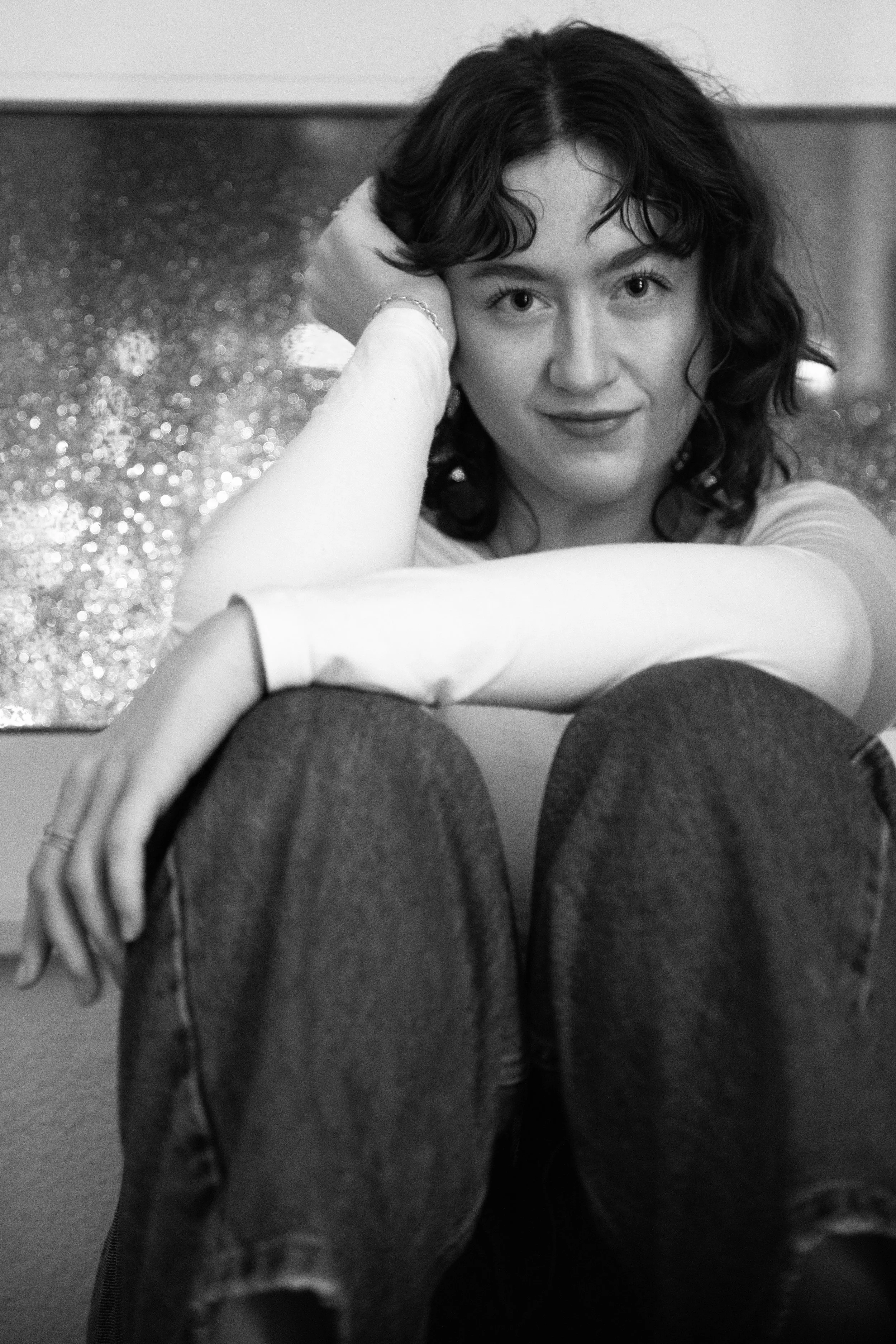 Une femme avec des cheveux bouclés courts en noir et blanc, assise avec ses jambes croisées, portant un haut clair et des jeans foncés, regardant vers l'objectif, en arrière-plan une surface brillante et un mur neutre.