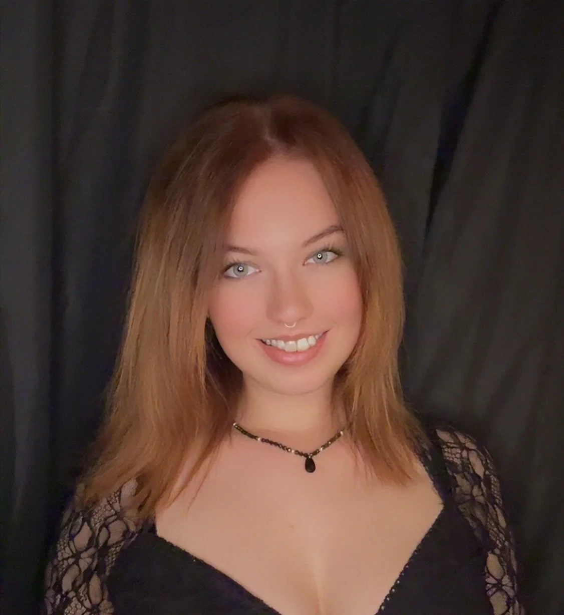 Ein junges Mädchen mit roten Haaren, blauen Augen und einem Lächeln, trägt einen schwarzen Spitzen-Top und eine schwarze Halskette, vor einem dunklen Hintergrund.