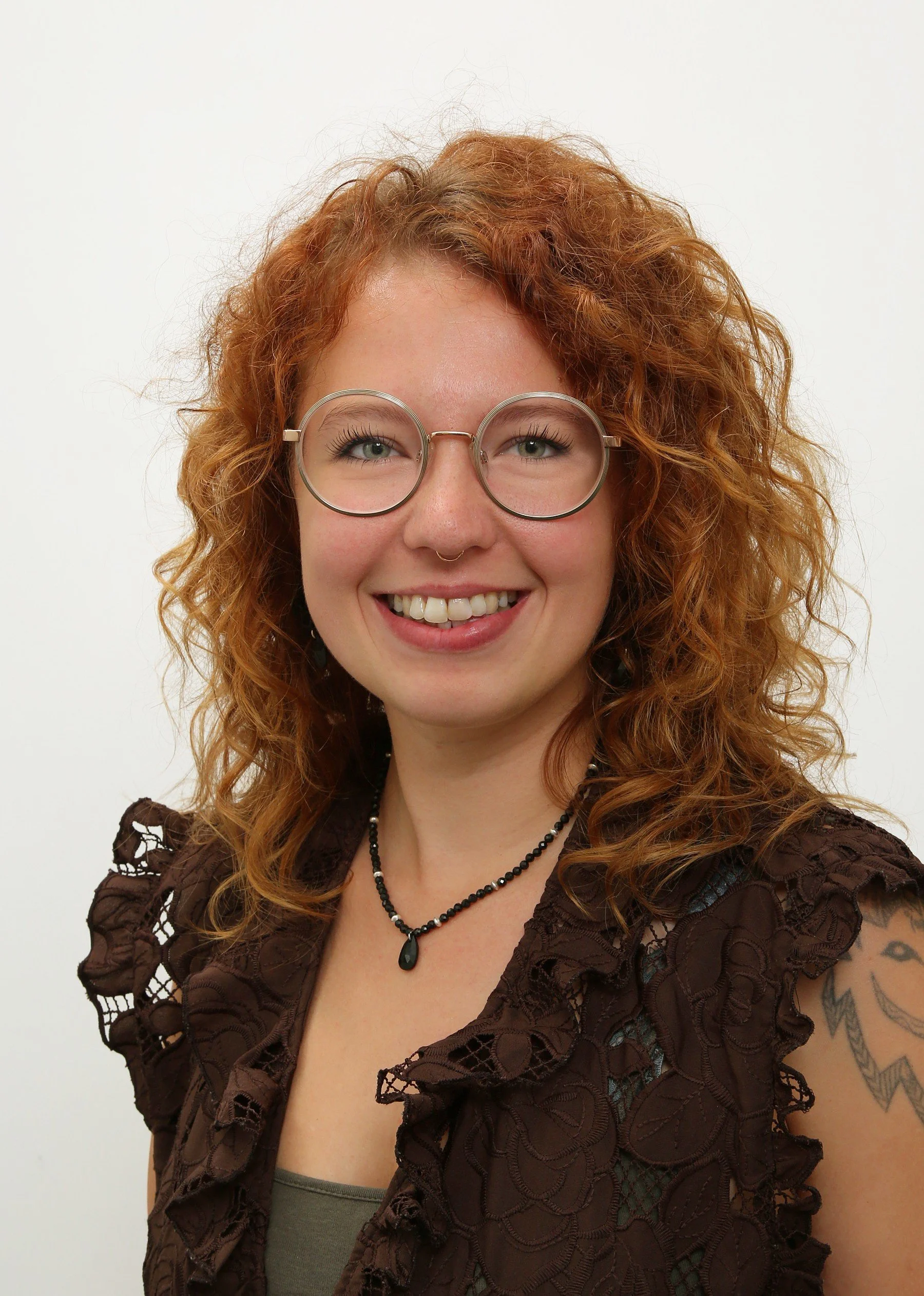 Porträt einer jungen Frau mit lockigem roten Haar und Brille, lächelnd vor weißem Hintergrund.