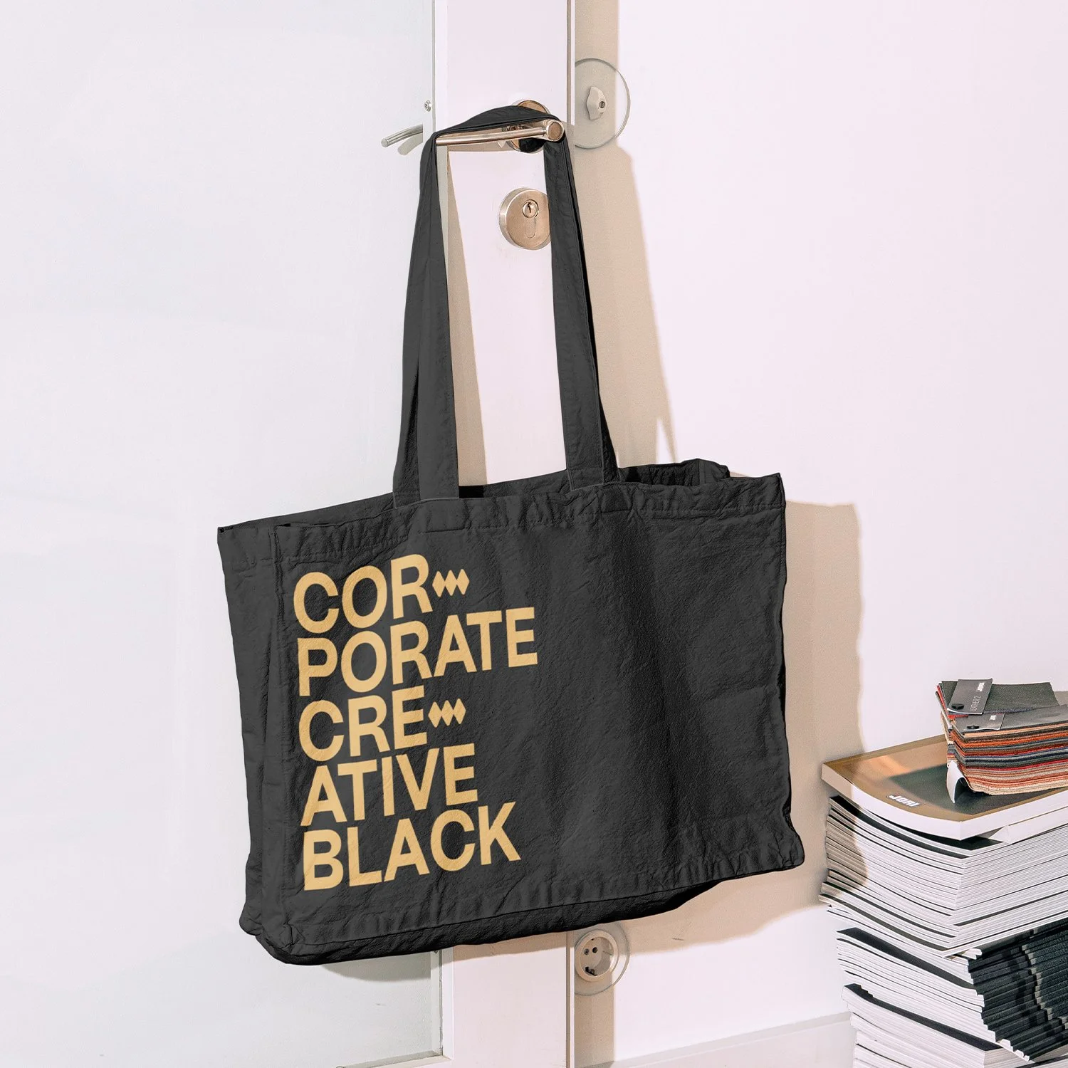 Black tote bag hanging on door handle with yellow text reading 'CORPORATE CREATIVE BLACK'.