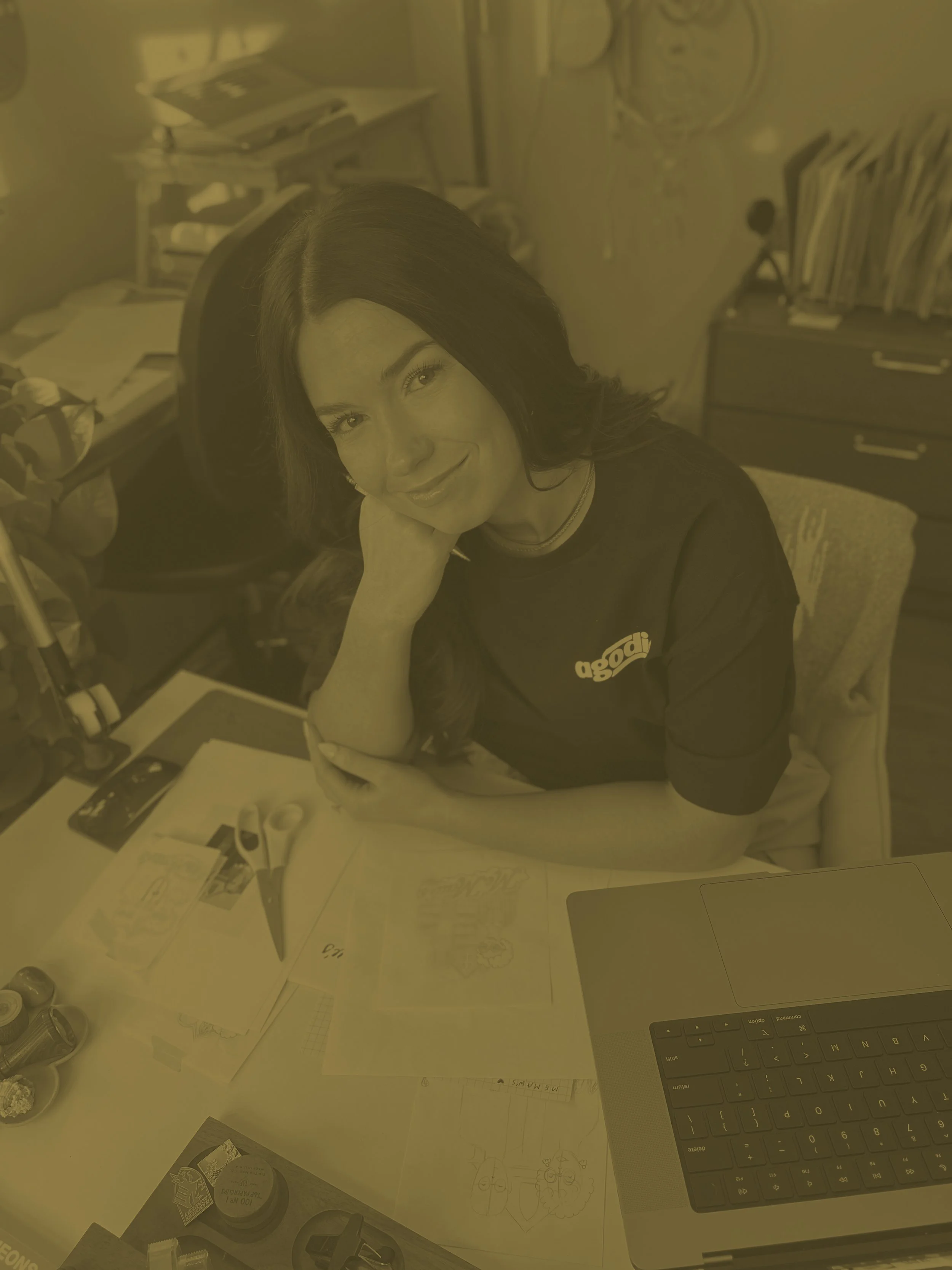 Desk with papers, drawing sketches, a laptop, and various office supplies. A woman with long dark hair, smiling, resting her chin on her hand, sitting at the desk.