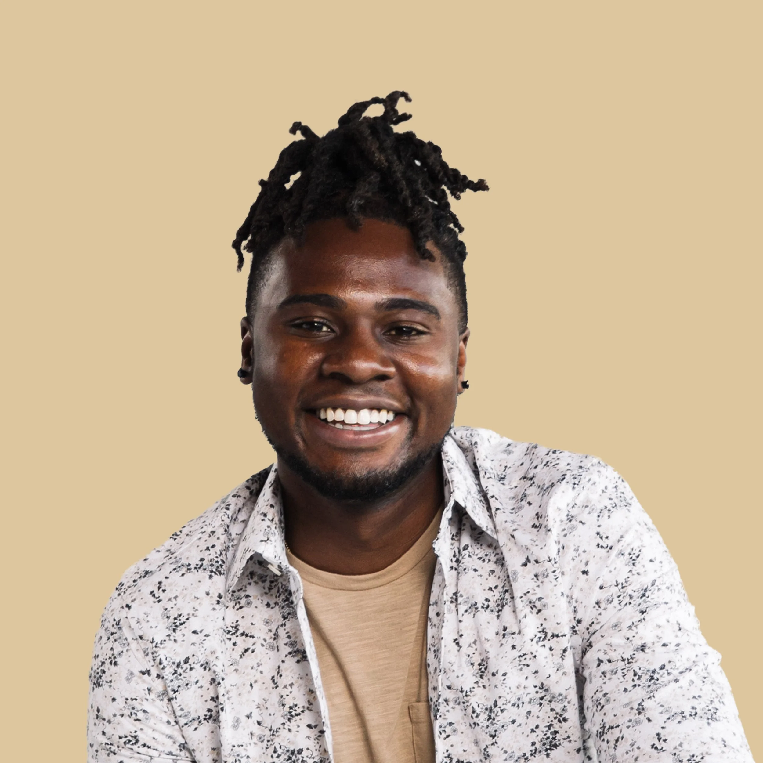 A person with dark skin and dreadlocks smiling at the camera, wearing a light-colored patterned shirt over a beige t-shirt, against a plain beige background.
