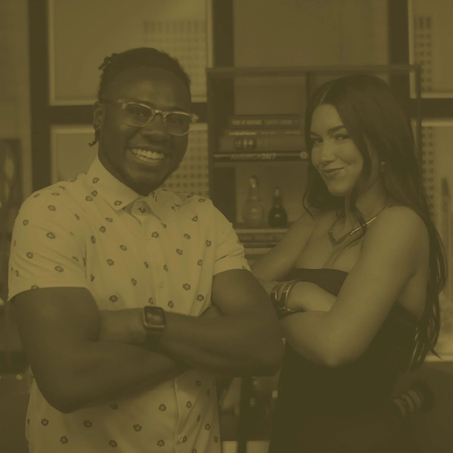 A man and woman smiling, sitting close together at a table in a bar or restaurant. The man is wearing glasses and a patterned shirt, the woman has long dark hair and is dressed in a sleeveless top. The background has shelves with books and bottles, suggesting an indoor casual setting.