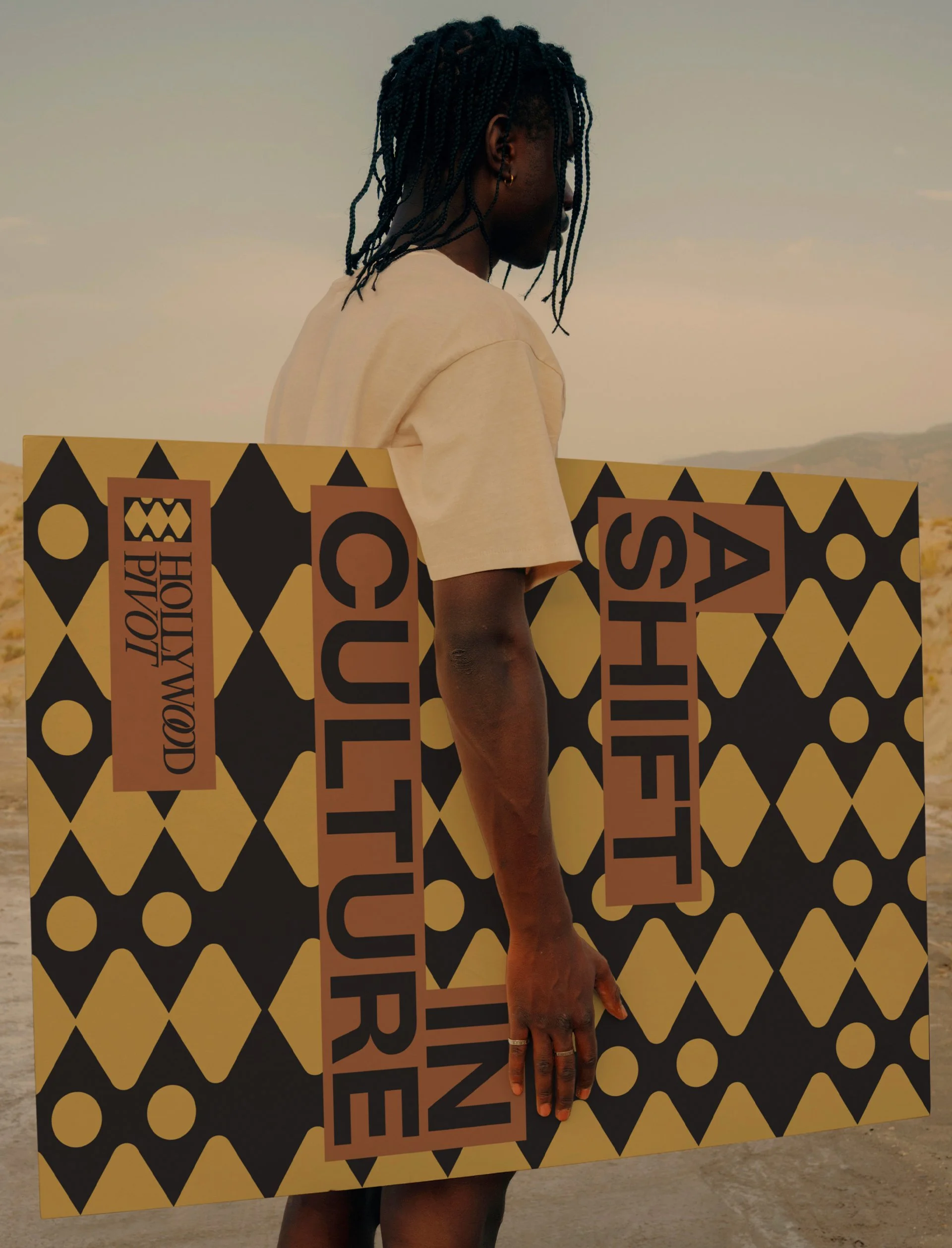 Person with braided hair wearing a beige shirt holding a large yellow sign with black geometric shapes and text, standing outdoors in a desert landscape under a cloudy sky.