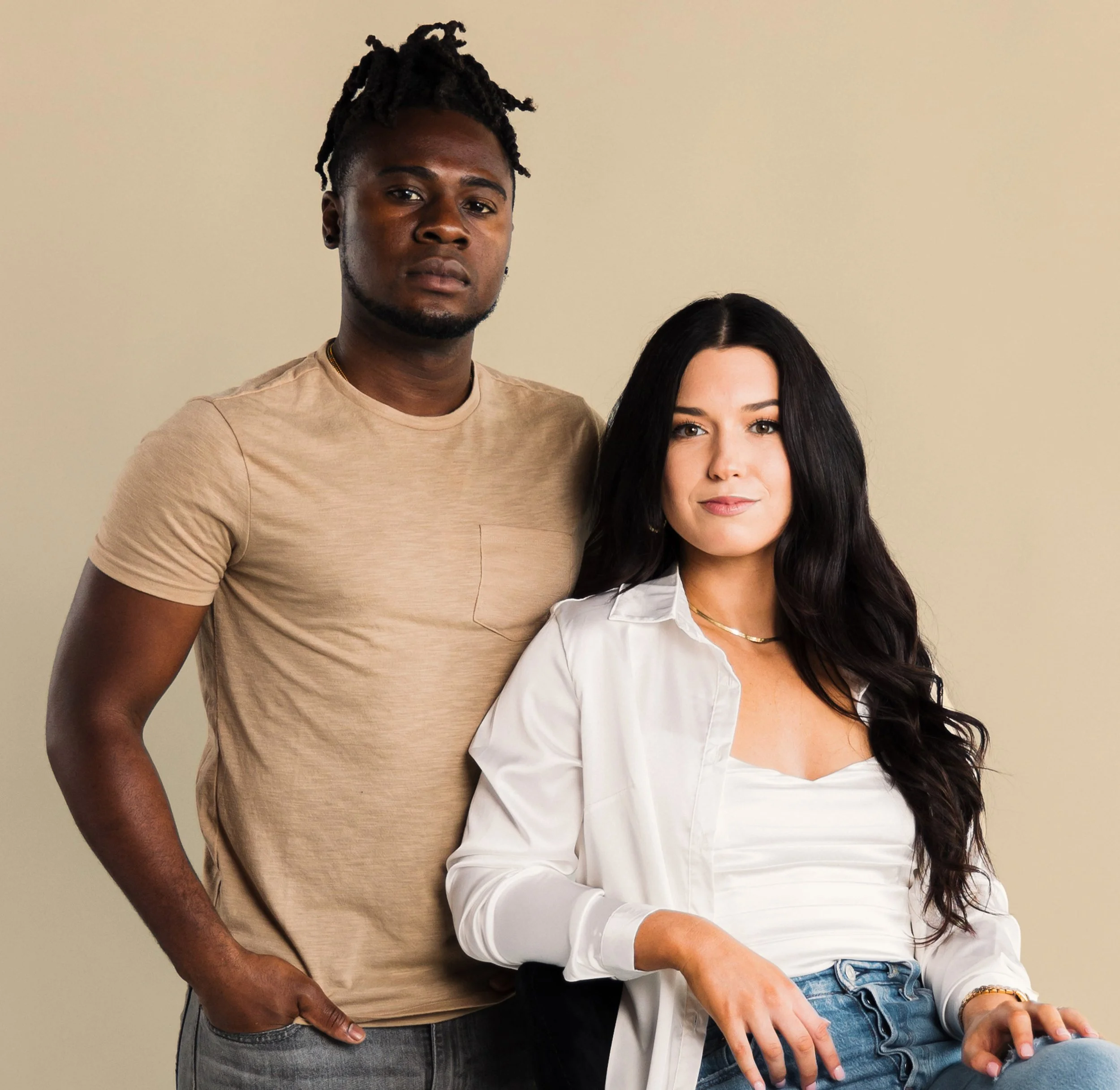 Close-up of a young man with dreadlocks and a woman with long dark hair posing against a plain background.