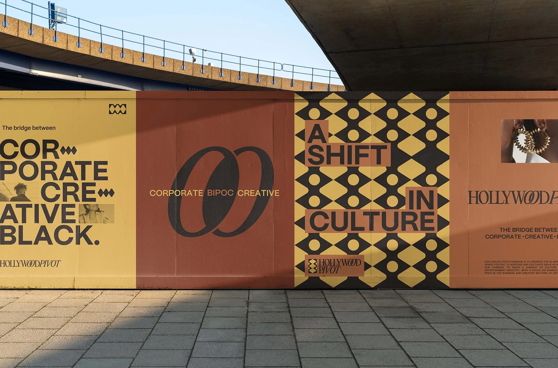 A colorful outdoor poster under a bridge promoting a creative and cultural event. The poster has bold black text on a yellow, orange, and black patterned background, with phrases like 'The bridge between corporate, BIPOC, creative' and 'A shift in culture.' It also mentions Hollywood Pivot and features a partial image of a person's head with large earrings.