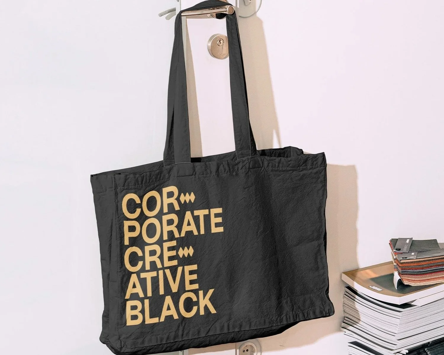 A black tote bag with beige text that reads 'Corporate Creative Black' hanging on a wall hook, next to a stack of magazines or catalogs on a small table.
