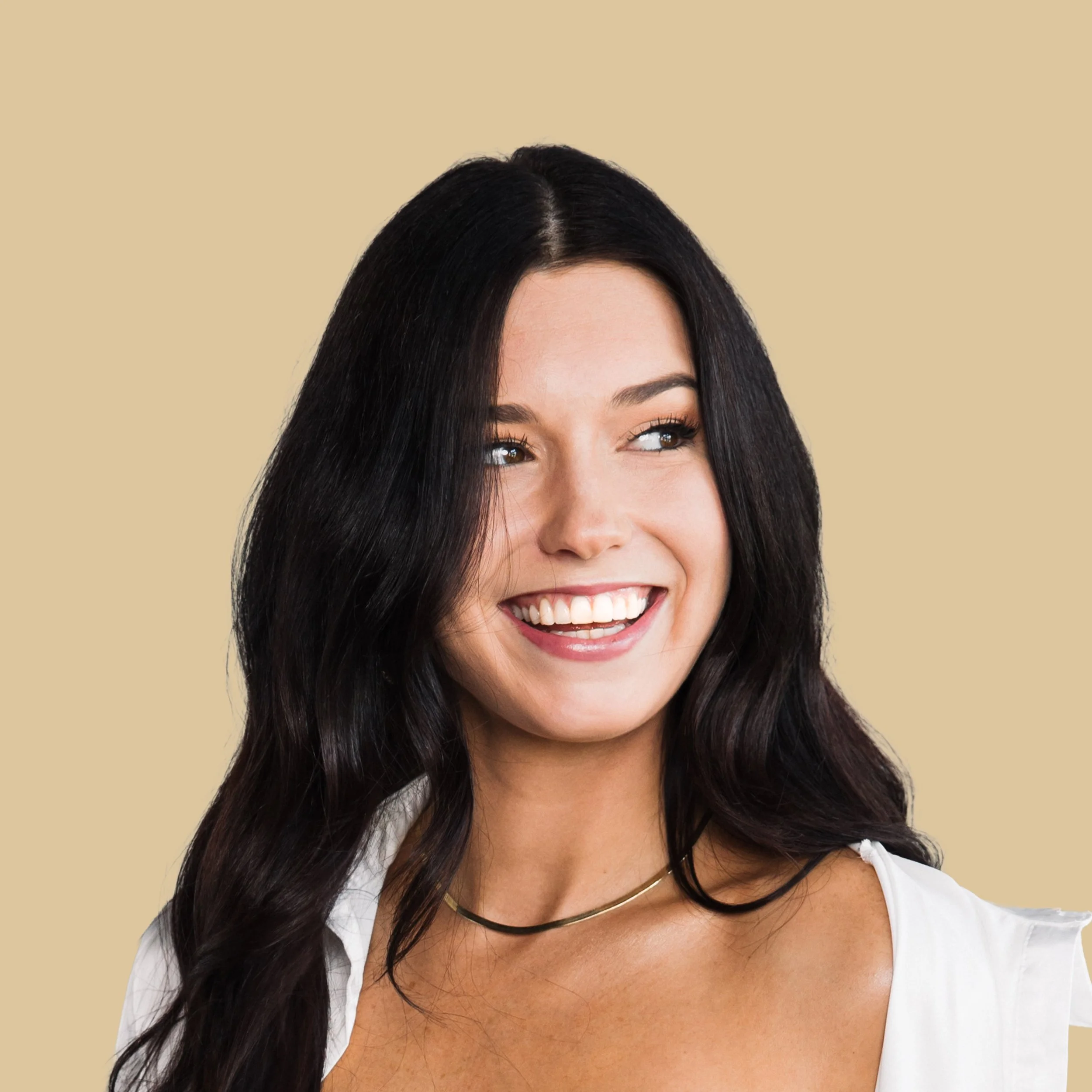Close-up of a smiling woman with long dark hair, wearing a white top and gold necklaces, against a beige background.