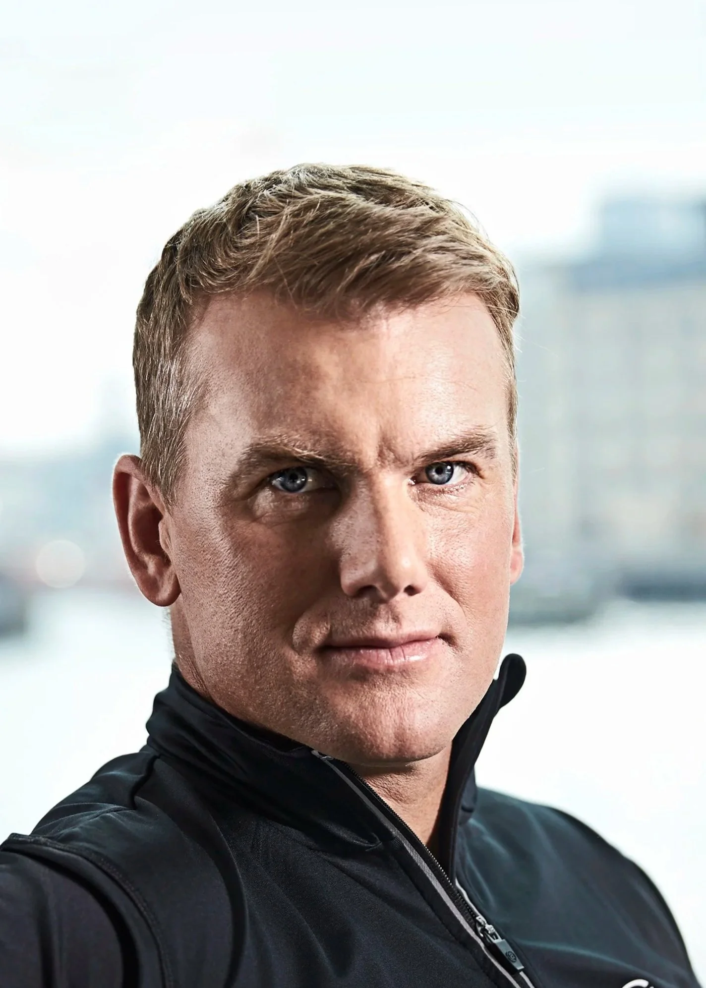 Close-up of a man with short light brown hair and blue eyes, wearing a black jacket with a zipper, outdoors with blurred cityscape background.