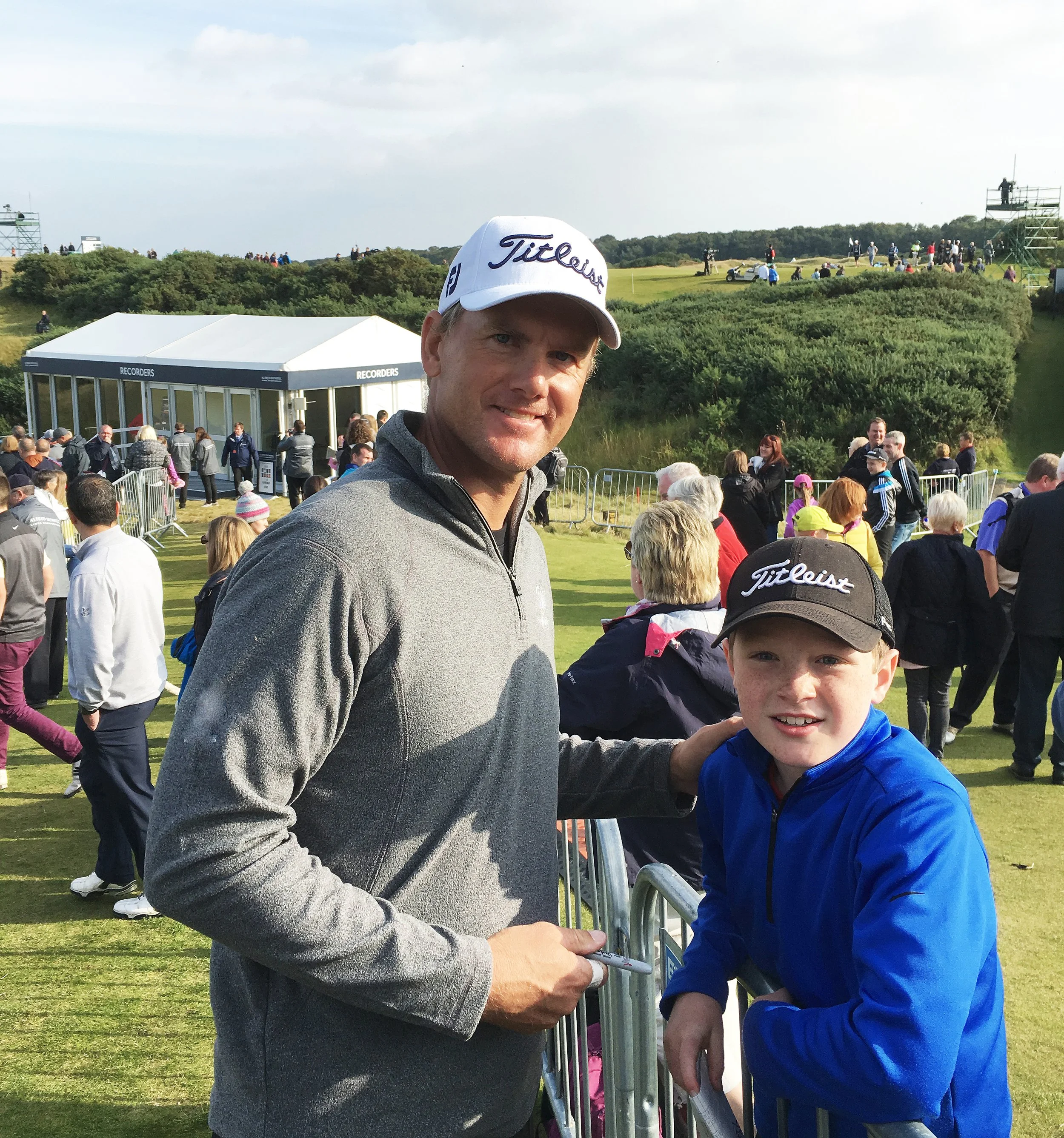 A man and a boy are at a golf tournament, signing an autograph at a barrier. The man is wearing a white Titleist cap and gray zip-up jacket. The boy is wearing a black Titleist cap and blue jacket. The background shows many spectators, a golf course, and a green hillside with scattered trees.