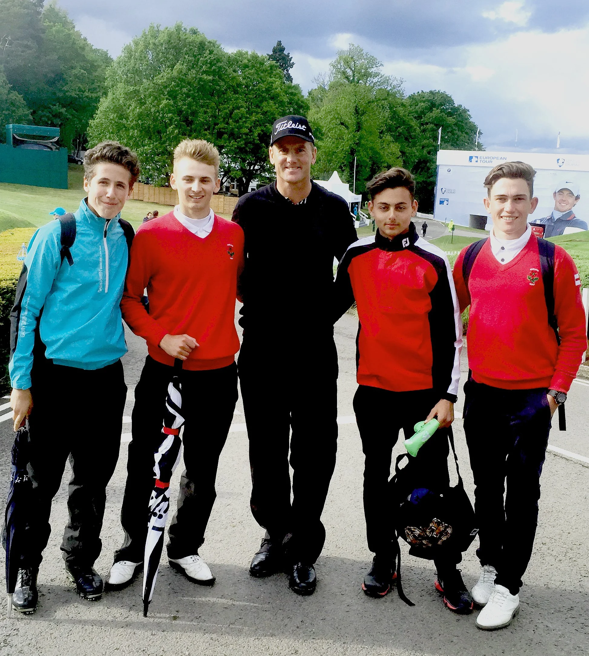 Group of five young men standing outdoors on a golf course, with trees and a tent in the background, dressed in sporty clothing.