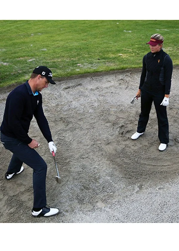 Two golfers practice bunker shots on a golf course, standing in a sand trap with golf clubs.