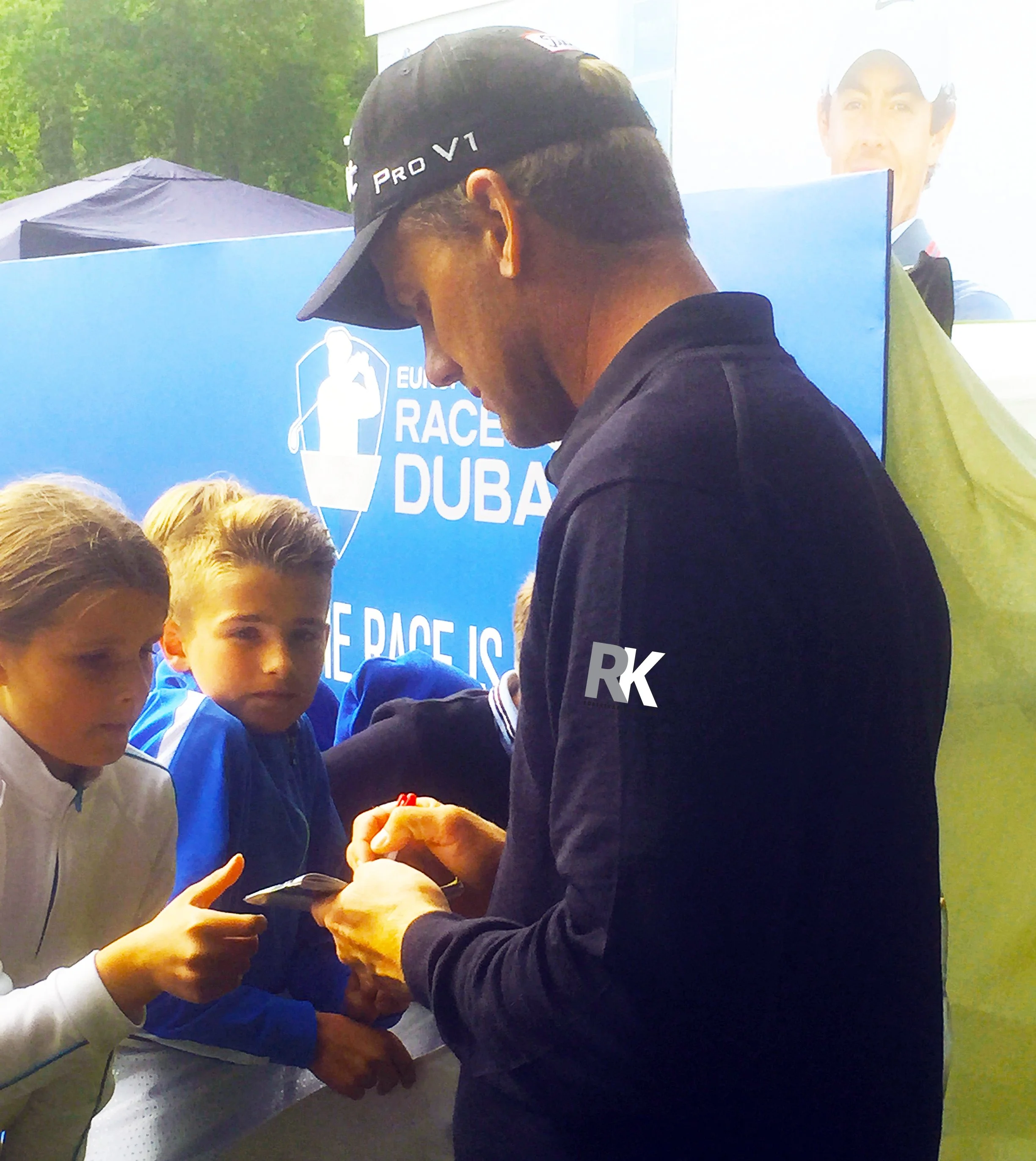 A man in a black cap receiving an autograph from a young boy in a blue jacket during a golf event, with a blue banner displaying the words 'Race Dubai' in the background.
