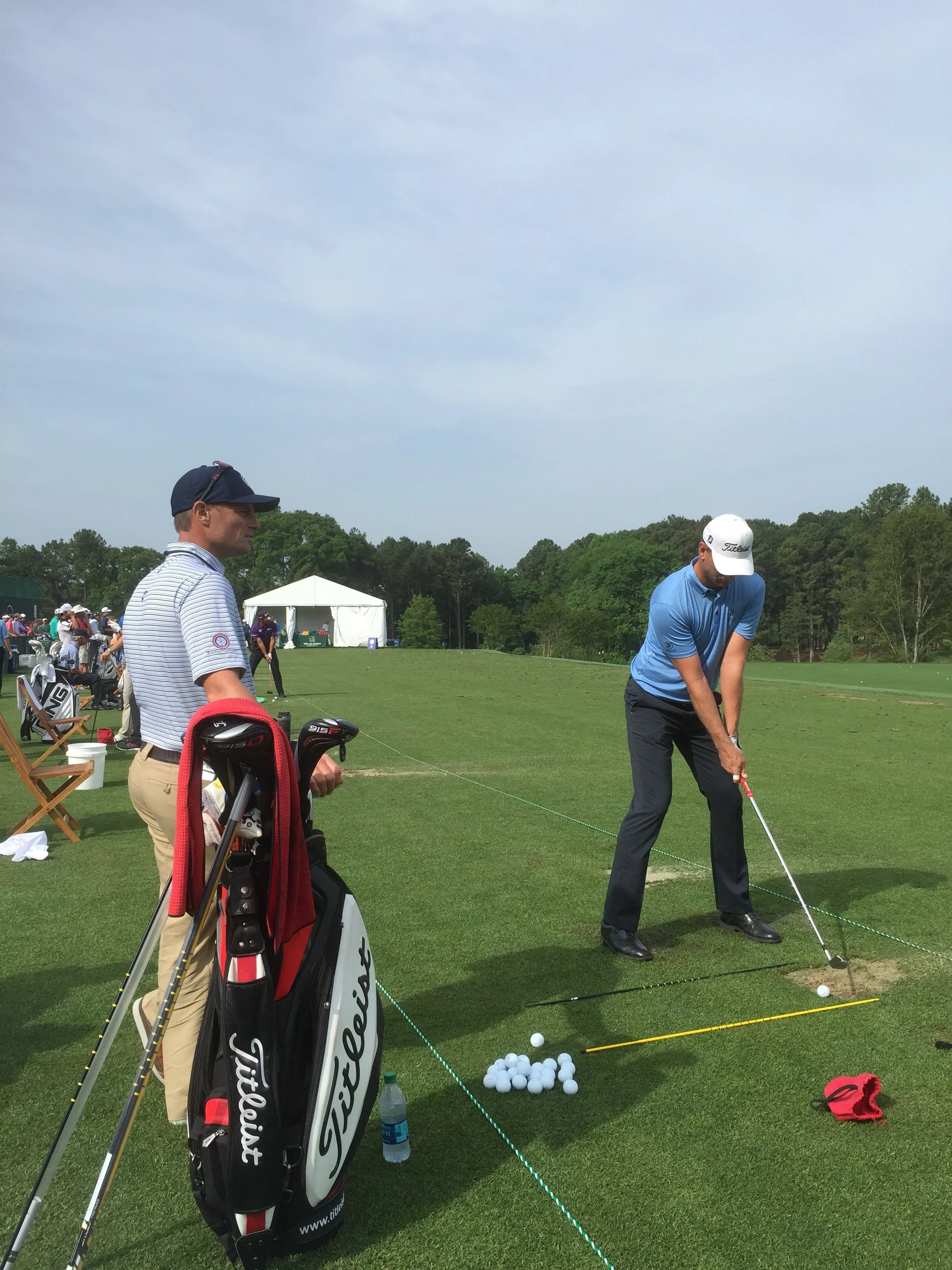 Two men practicing golf on a driving range, one is standing with golf clubs and the other is hitting a ball. There are golf balls on the ground, a golf bag, and a tent in the background.